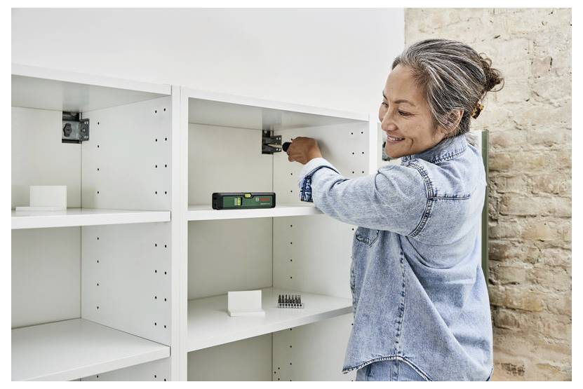 Une personne souriante en train de monter une unité de rangement blanche contre un mur de briques, utilisant des outils.