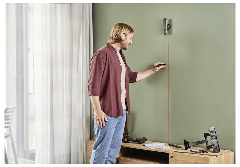 Une personne utilise un niveau laser pour tracer une ligne verticale sur un mur vert. Un perceuse et du matériel de montage sont disposés sur la table à proximité.
