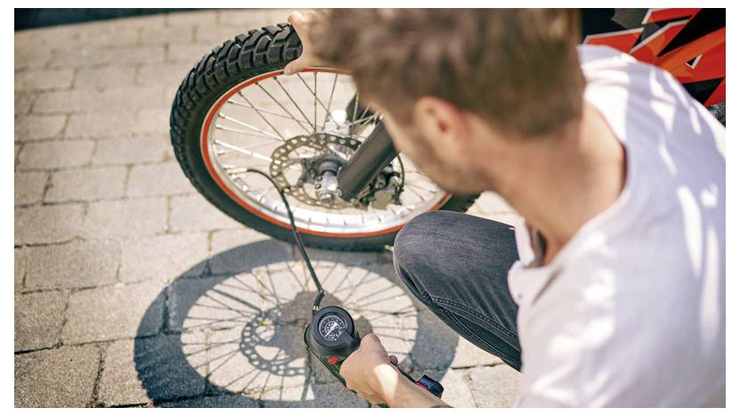 Une personne s'agenouille sur un chemin pavé, gonflant le pneu avant d'une moto à l'aide d'une pompe à air portable.
