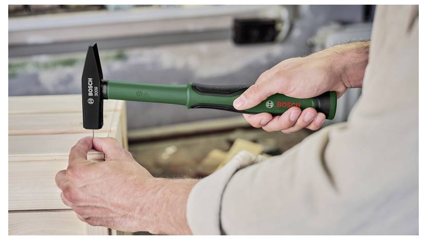 Une personne utilise un marteau Bosch vert et noir pour clouer un panneau en bois, illustrant une activité de rénovation domiciliaire ou de travail du bois.