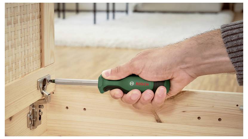 Une main d'une personne utilisant un tournevis vert pour visser une vis sur la charnière d'un meuble en bois, suggérant un travail de montage ou de réparation.