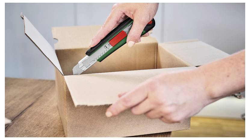 Une personne utilise un cutter pour ouvrir un carton sur une table en bois.