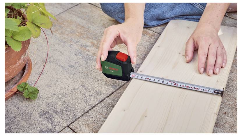 Une personne mesure une planche en bois à l'aide d'un mètre ruban sur une surface carrelée, à côté d'un pot de plante.