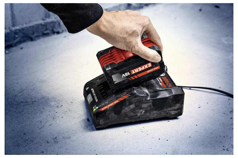 Une main plaçant une batterie rouge « Expert 18V » sur une station de charge noire sur une surface en béton, indiquant un processus de charge de batterie.