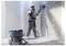 Un travailleur portant un équipement de protection ponce un mur dans une pièce en construction, utilisant une ponceuse électrique raccordée à un système d'aspiration.