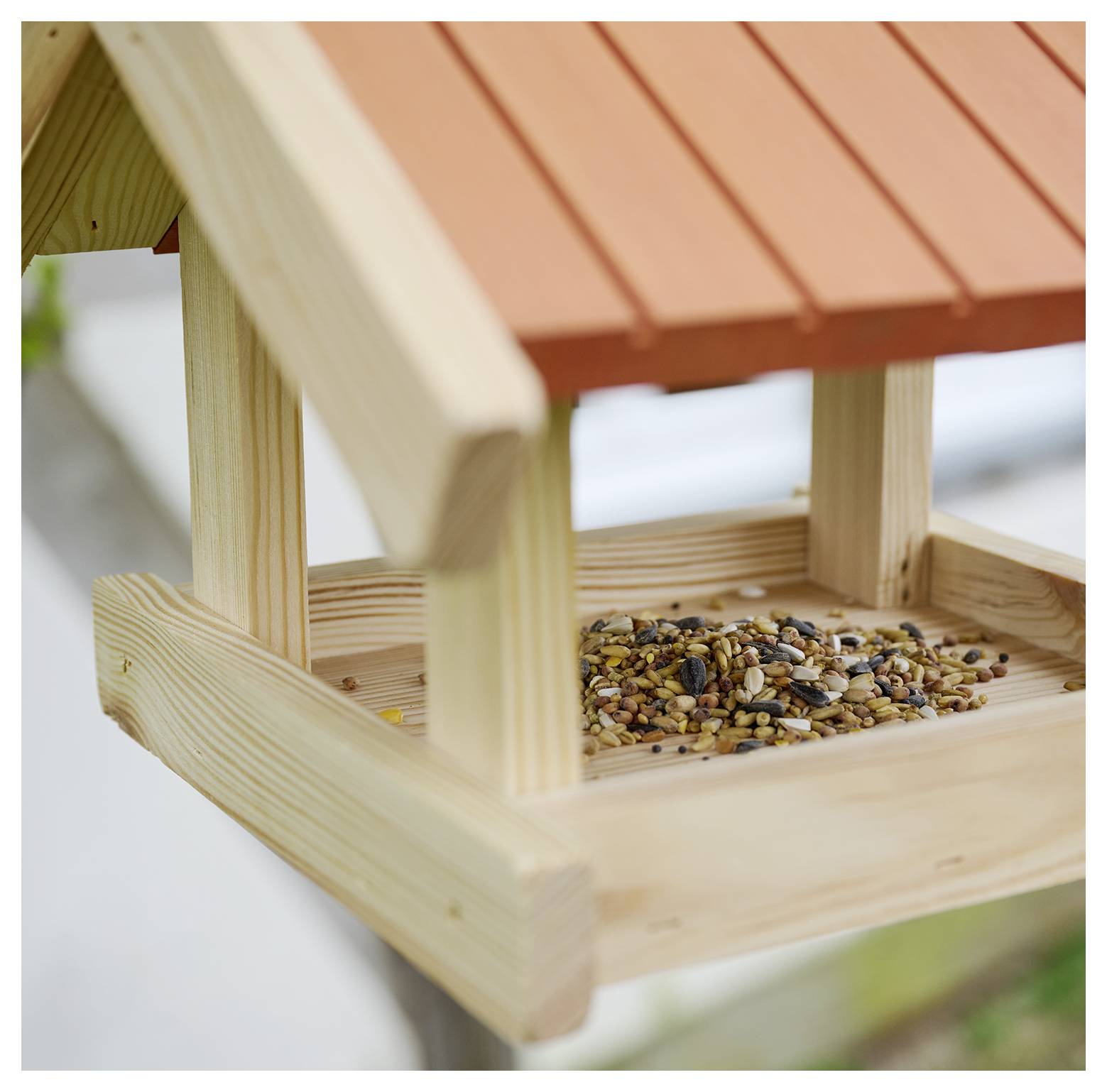 Un mangeoire en bois remplie de graines mélangées, située sous un toit orange, invite les oiseaux à se restaurer dans un cadre de jardin.