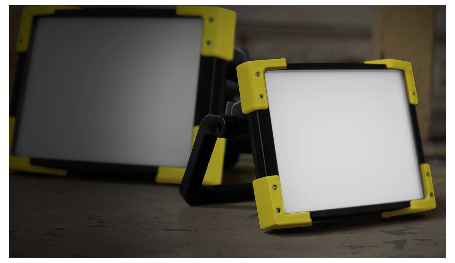 Deux projecteurs de travail portables à LED avec des châssis jaunes sur un sol industriel, l'un orienté vers l'avant et l'autre incliné sur le côté.