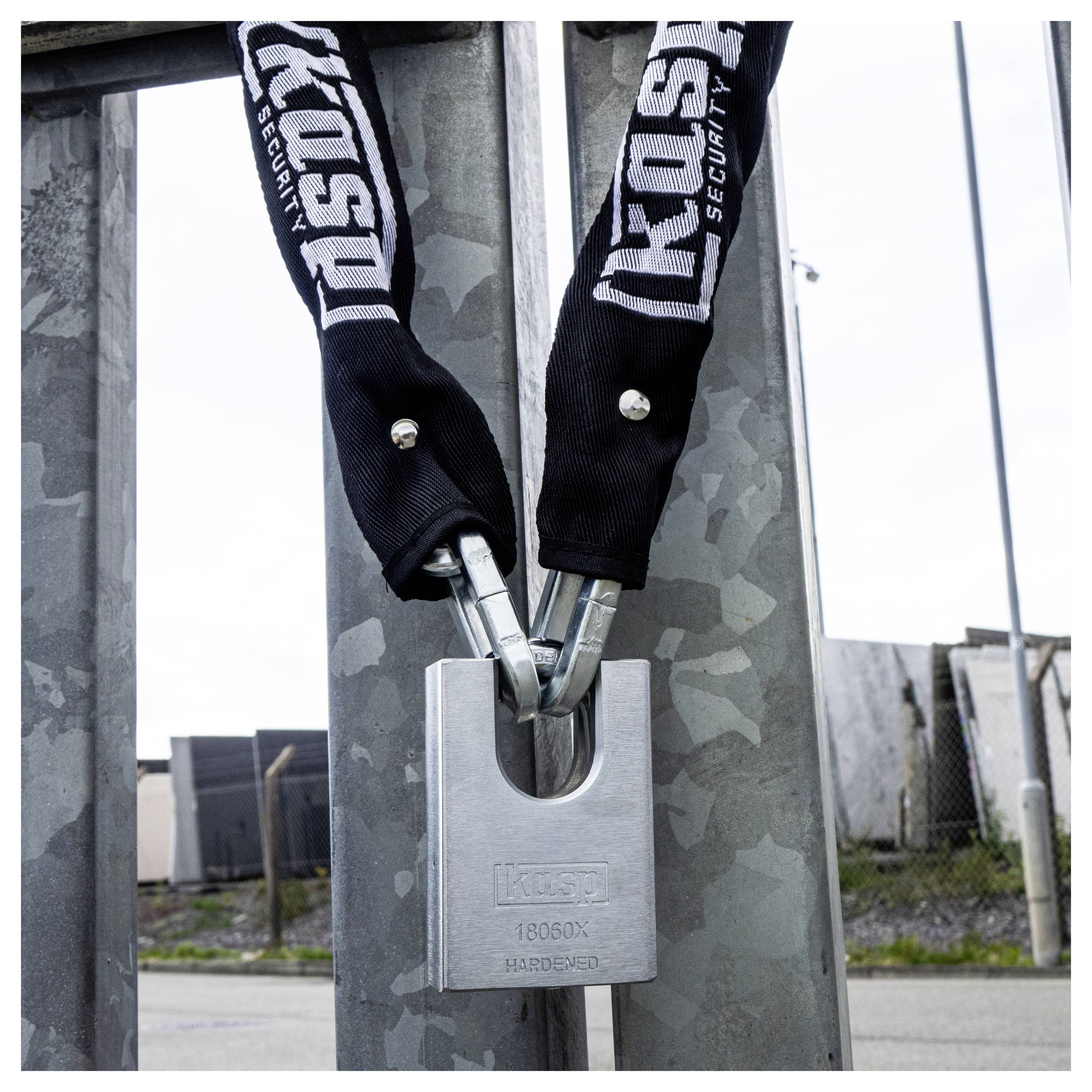 Un cadenas argenté sécurisant une chaîne portant l'inscription « KOSU SECURITY » imprimée sur un tissu noir, attaché à un portail métallique ; arrière-plan industriel.