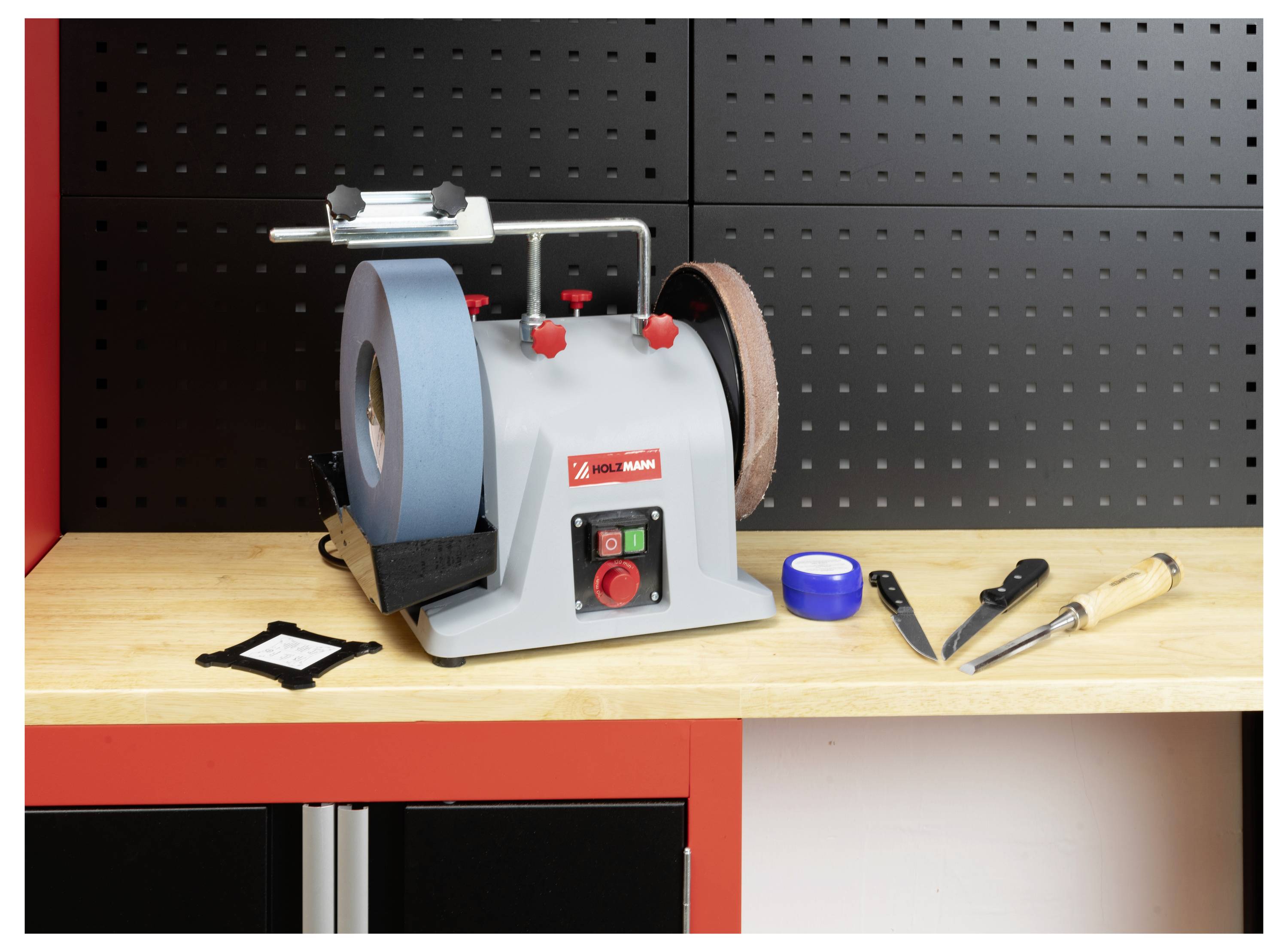 Un touret d'établi équipé d'une meule de affûtage bleue et d'une meule de polissage brune est posé sur un établi en bois. À proximité se trouvent un ciseau, un couteau et du ruban adhésif bleu.