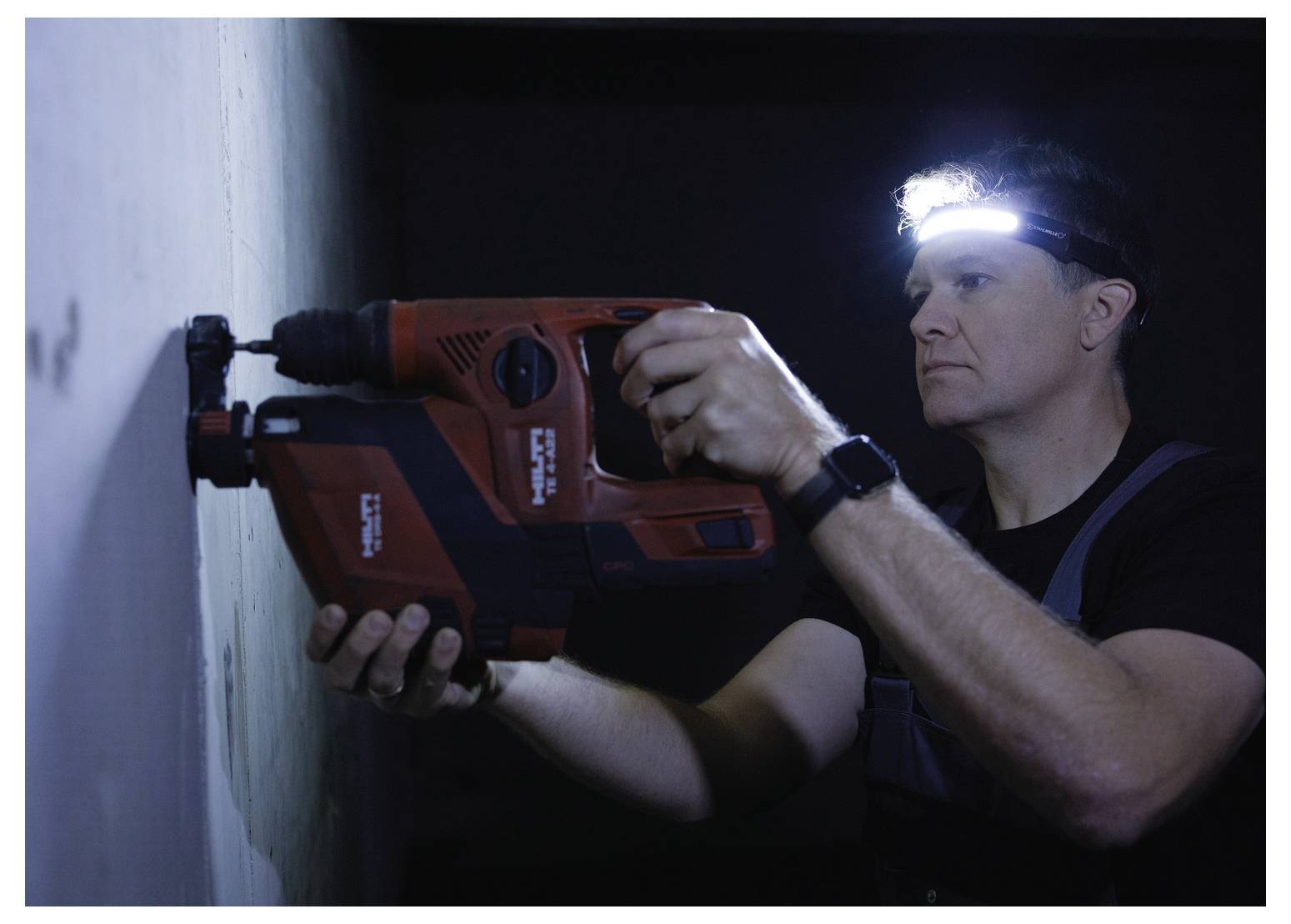 Une personne portant une lampe frontale utilise une perceuse électrique contre un mur dans un environnement faiblement éclairé, concentrée avec intensité sur sa tâche.
