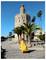 Une femme en robe jaune fluide marche près de la Torre del Oro avec des palmiers et un ciel bleu clair en arrière-plan.
