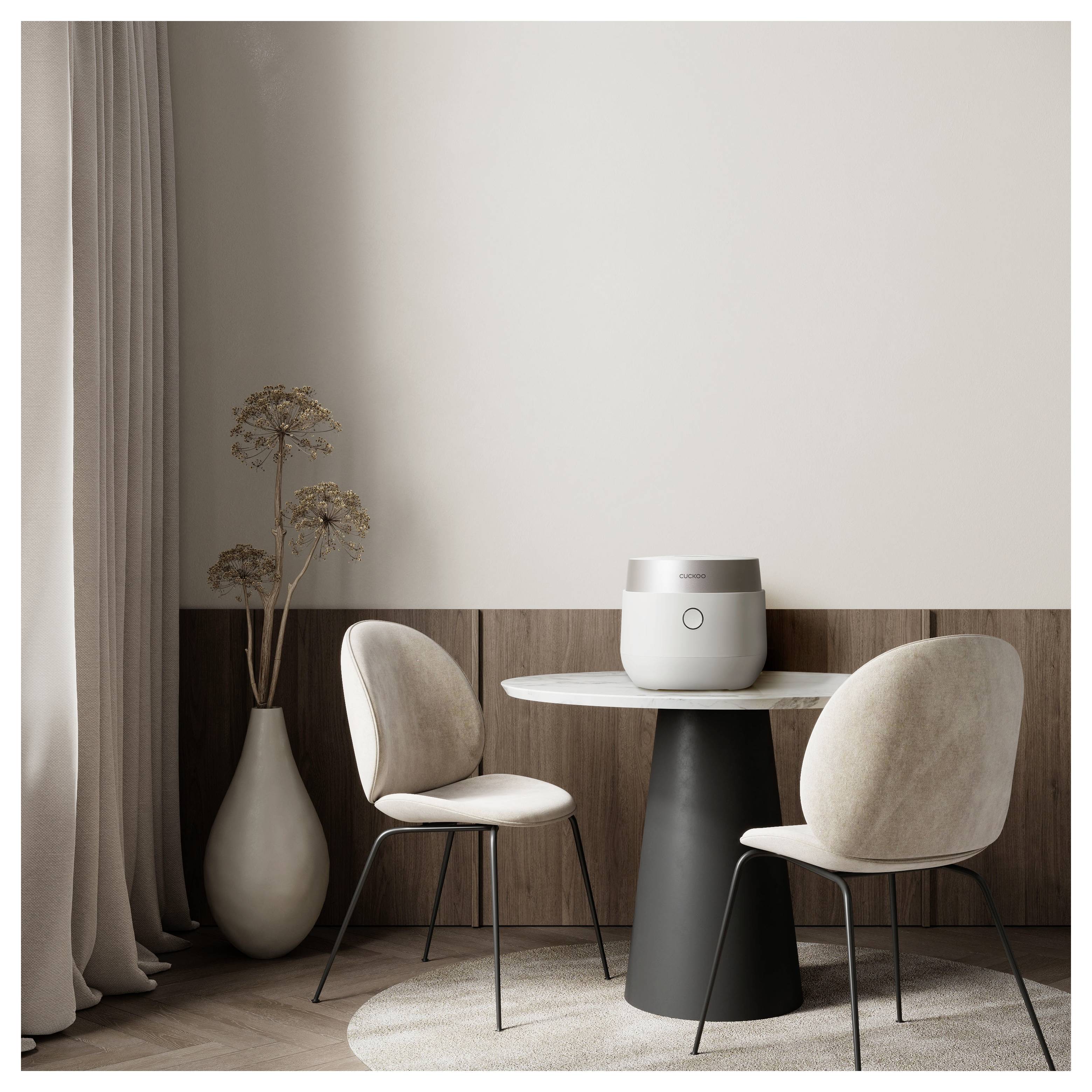 Un espace salle à manger minimaliste avec une petite table ronde, deux chaises beiges, une plante en pot et un appareil rond posé sur la table, contre un mur neutre.