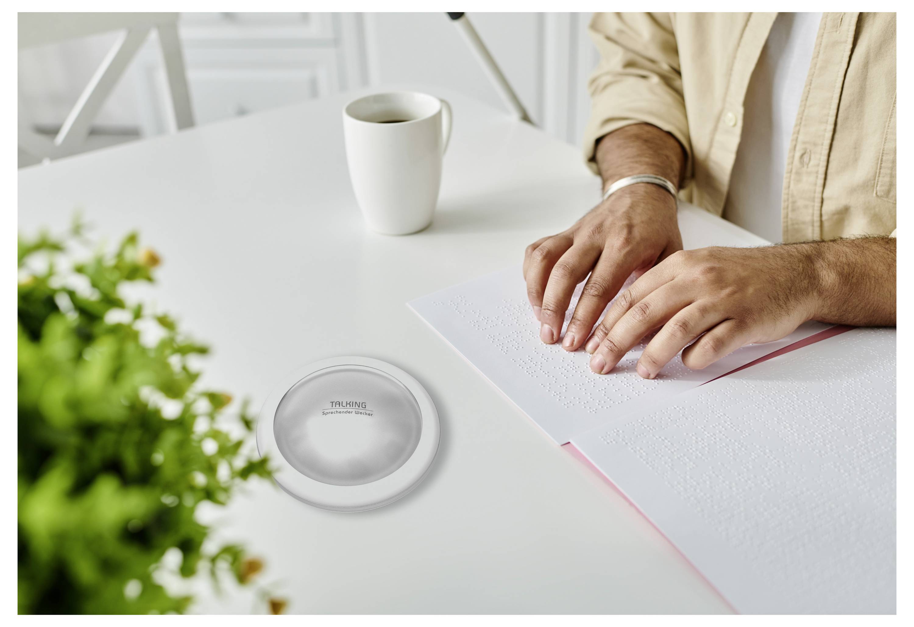 Une personne lit du braille à une table blanche, avec une tasse et une petite plante à proximité, soulignant l'accessibilité et l'apprentissage tactile.