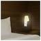 Une lampe beige à faible éclairage est fixée à un mur en bois sombre au-dessus d'un oreiller blanc et de draps sur un lit, créant une atmosphère calme et chaleureuse.