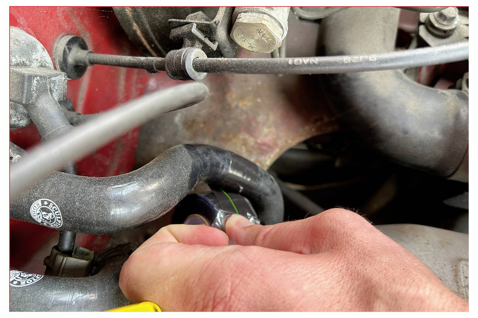 Une main tournant un bouchon noir sur un tuyau du moteur d'un véhicule, entourée de divers tuyaux connectés et de pièces métalliques, indiquant un entretien.