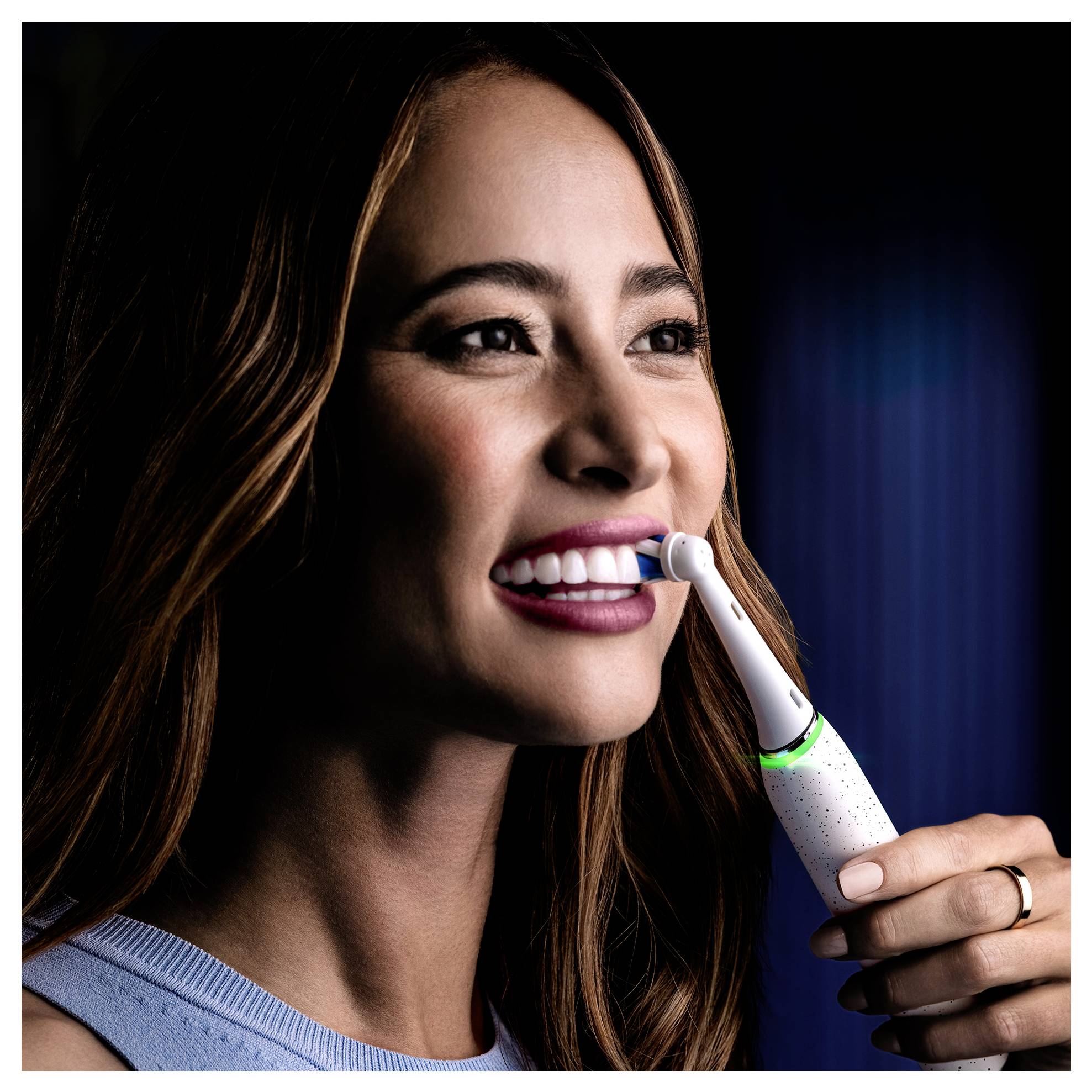 Une personne aux cheveux longs utilise une brosse à dents électrique, souriante, démontrant une bonne hygiène bucco-dentaire. La brosse à dents possède un indicateur lumineux en marche.