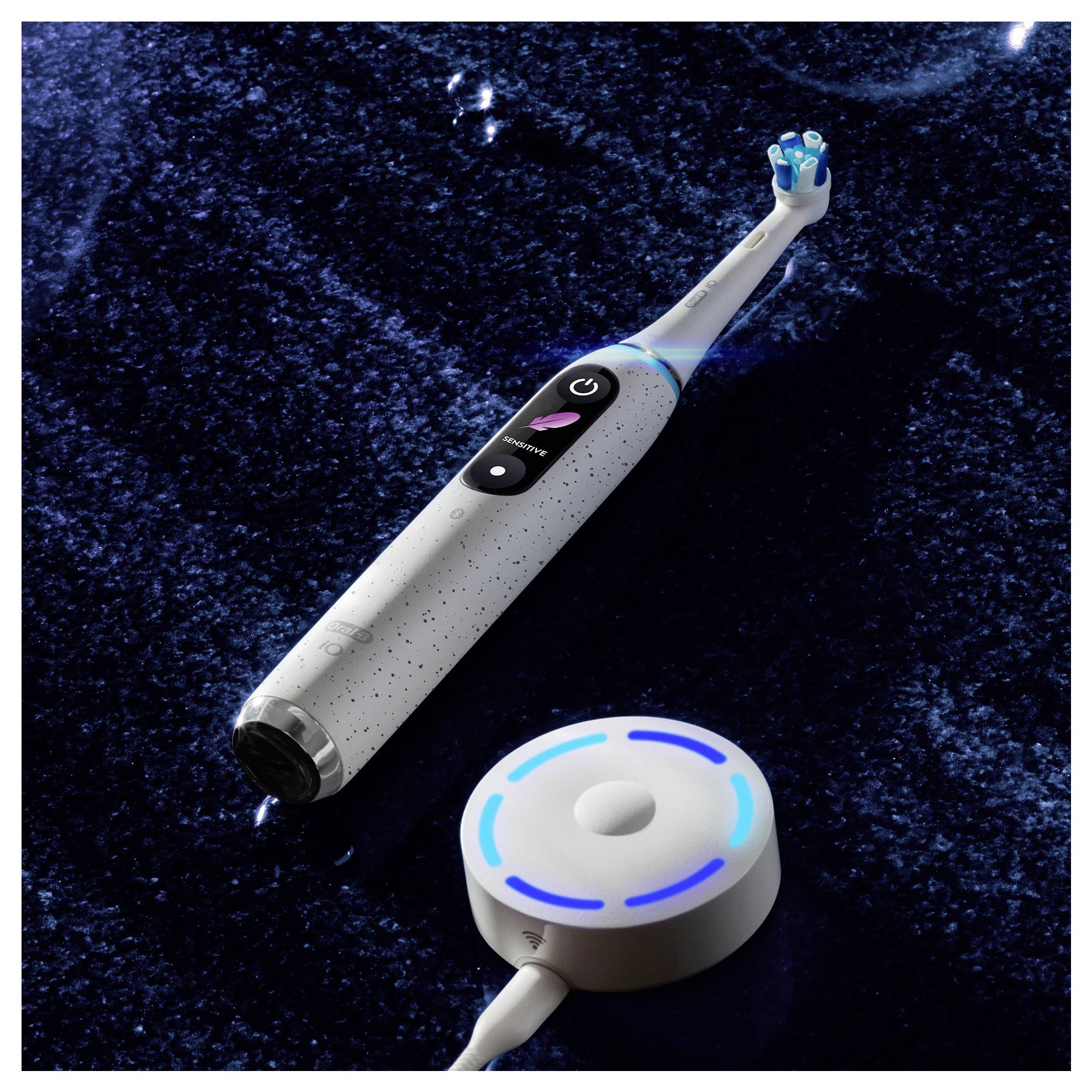 Une brosse à dents électrique avec un manche blanc et un affichage numérique est posée sur une surface brillante et sombre. Une base de chargement circulaire est à proximité, illuminée en bleu.