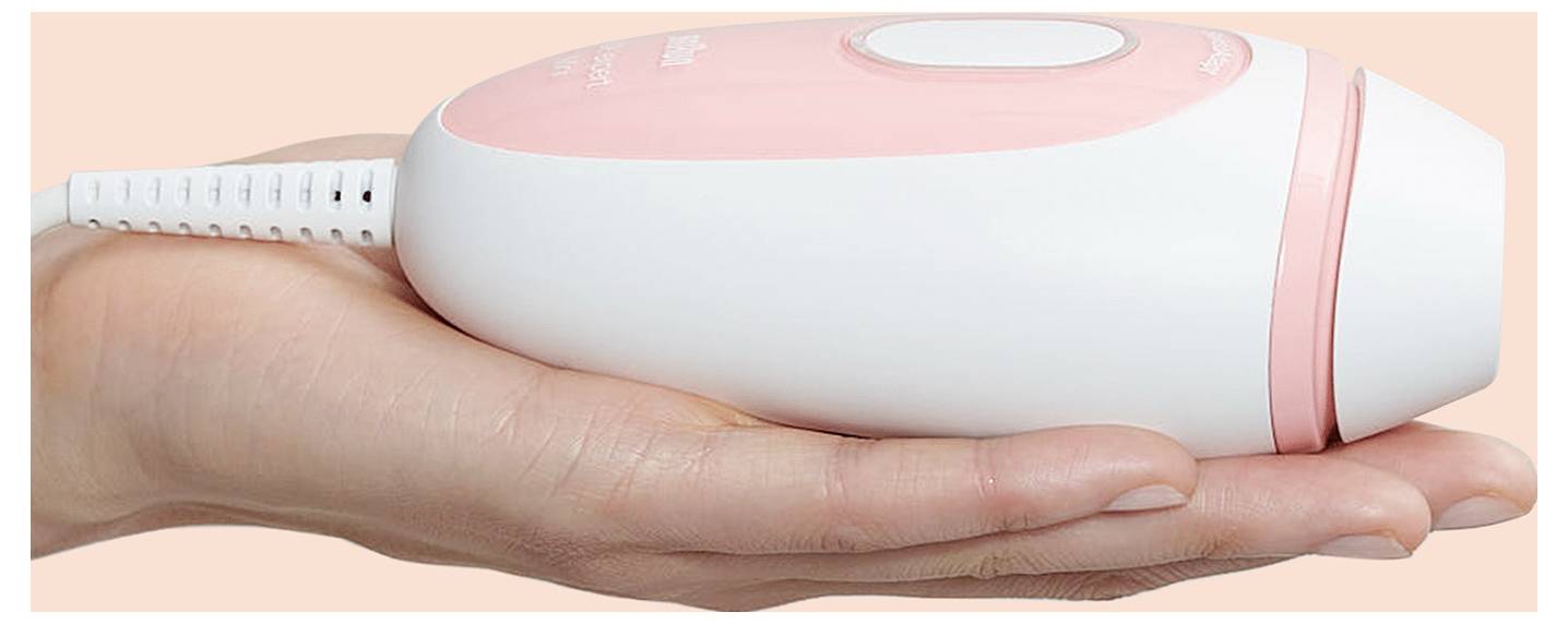 Une main tient un petit appareil électronique blanc et rose compact, avec un bouton sur le dessus, probablement un outil de soin personnel ou un dispositif de beauté.