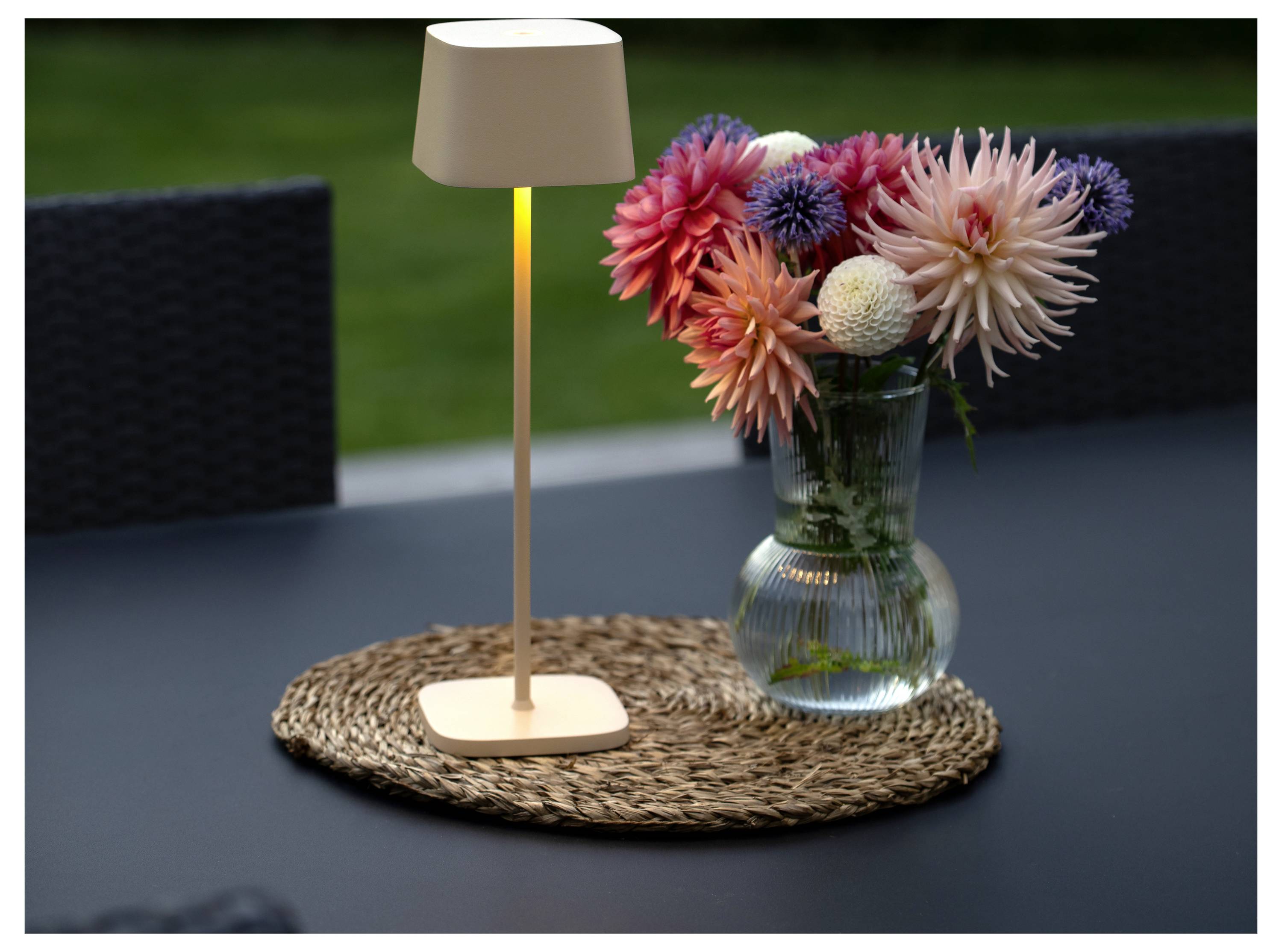 Une petite lampe de table allumée et un vase de fleurs colorées sur un tapis tressé posé sur un plateau sombre, créant une ambiance chaleureuse et extérieure.