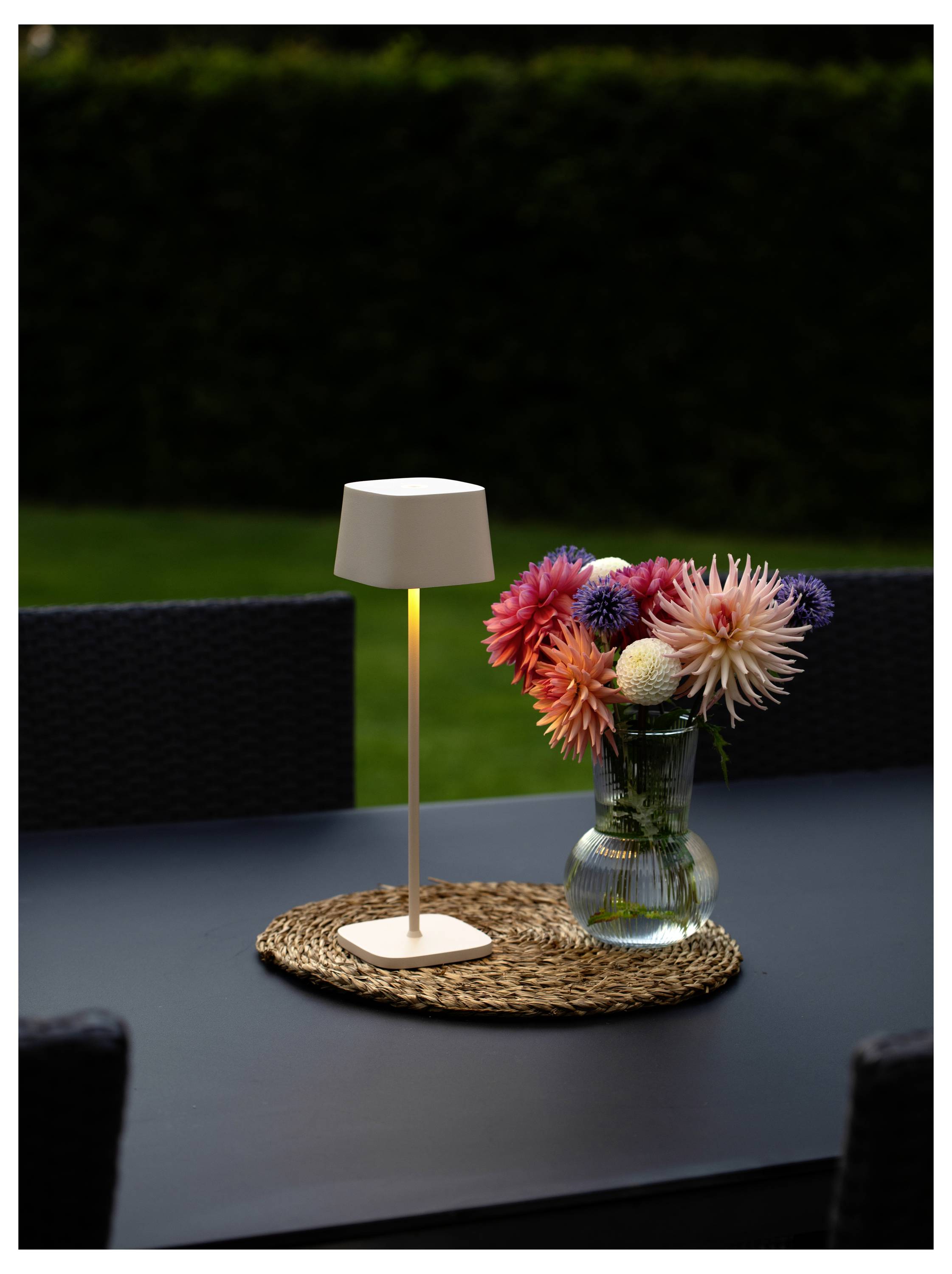 Un vase rempli de fleurs colorées et une lampe blanche moderne sont posés sur un tapis tissé, sur une table sombre à l'extérieur, avec un jardin flou en arrière-plan.