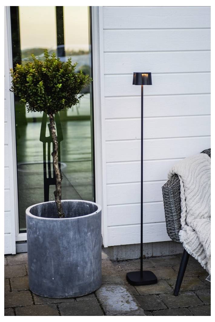 Un arbre en pot et une lampe élégante et droite à côté d'un fauteuil recouvert d'une couverture texturée, disposés devant un bardage blanc et une porte vitrée.