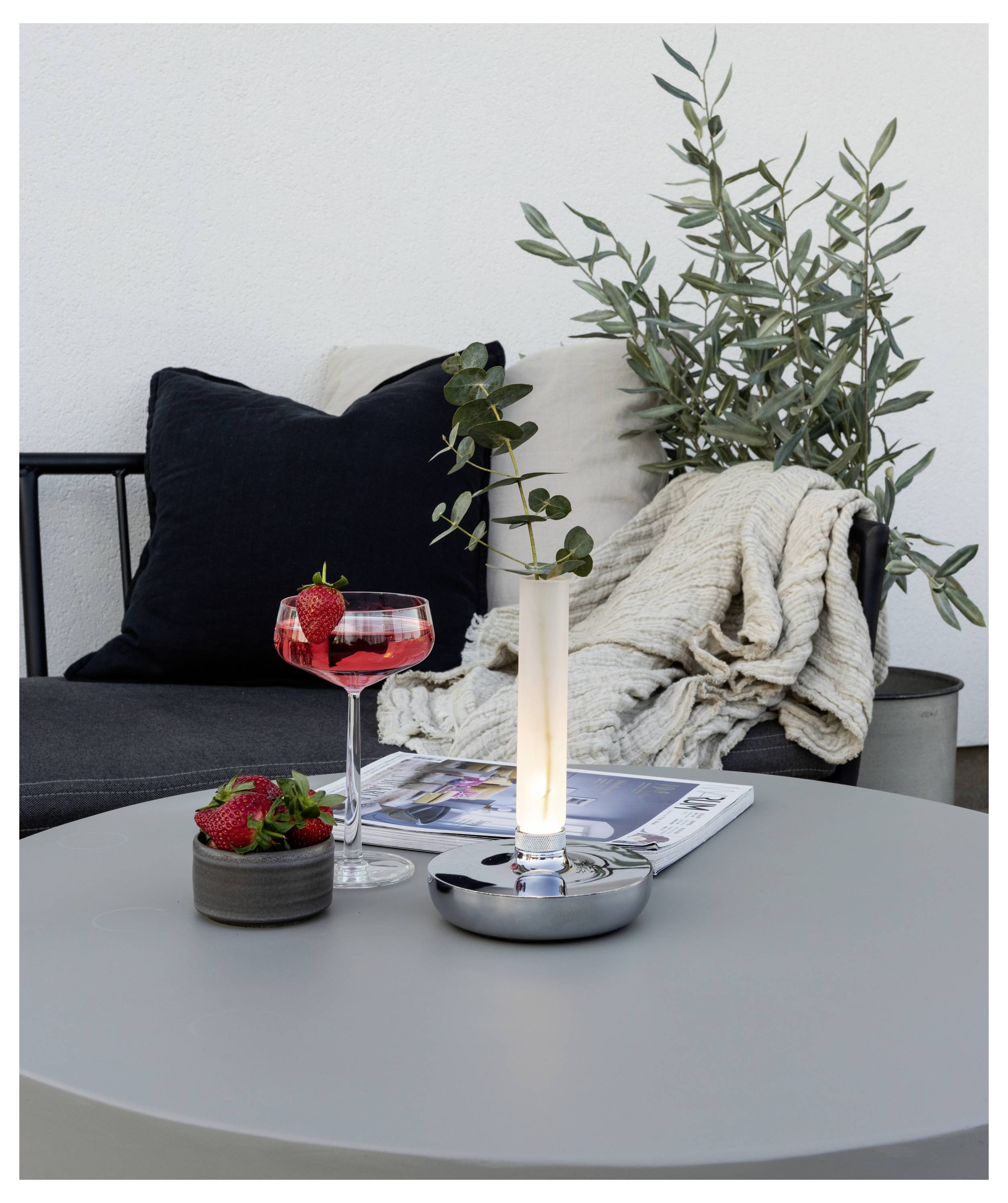 Un espace salon moderne avec une table grise sur laquelle sont disposés un cocktail garni de fraises, un bol de fraises, une bougie et un journal.