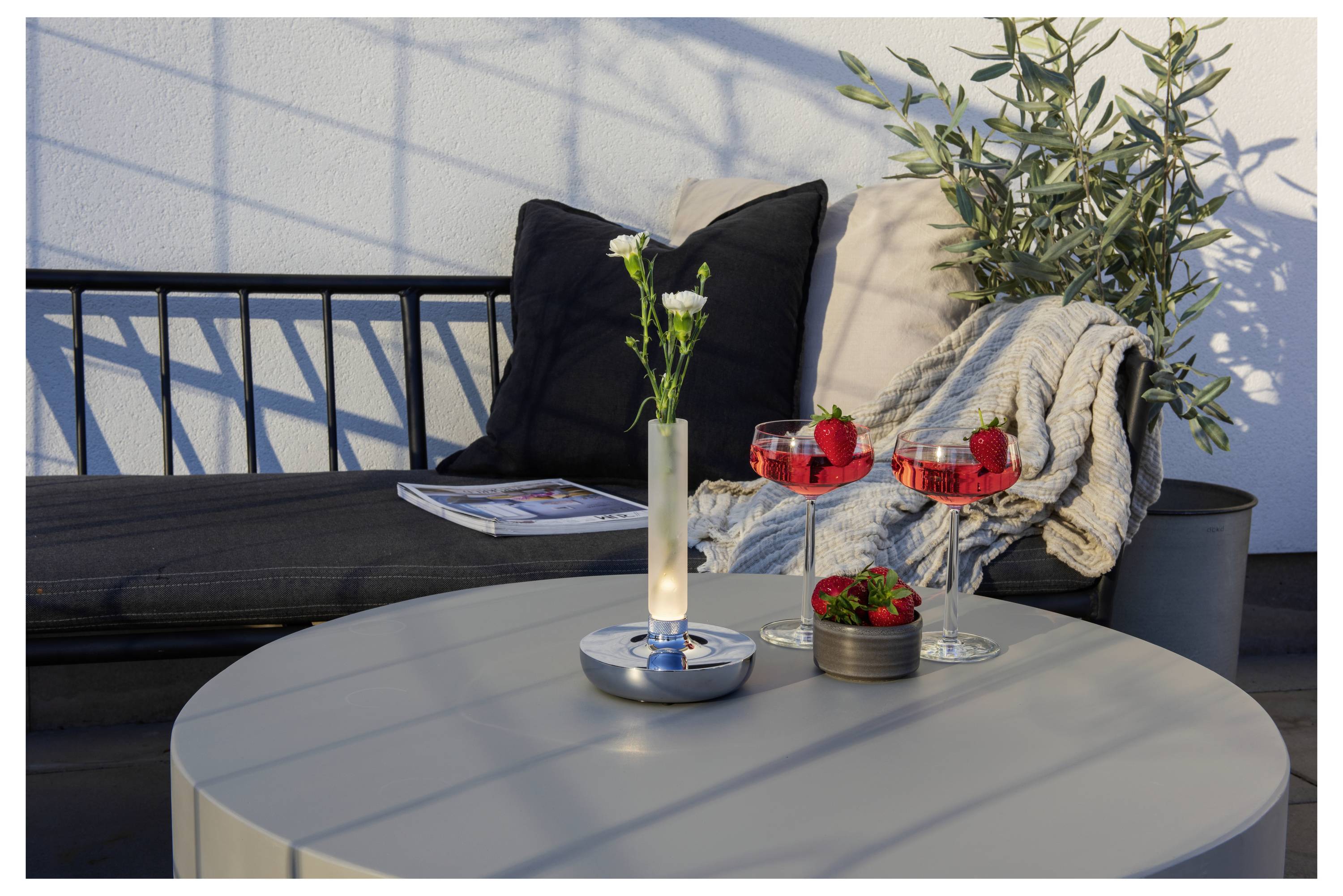 Un aménagement de patio élégant présente une table grise avec deux cocktails rouges ornés de fraises, un petit vase contenant des fleurs blanches, et un banc noir avec coussin, sur lequel est posée une couverture, avec une plante en arrière-plan.