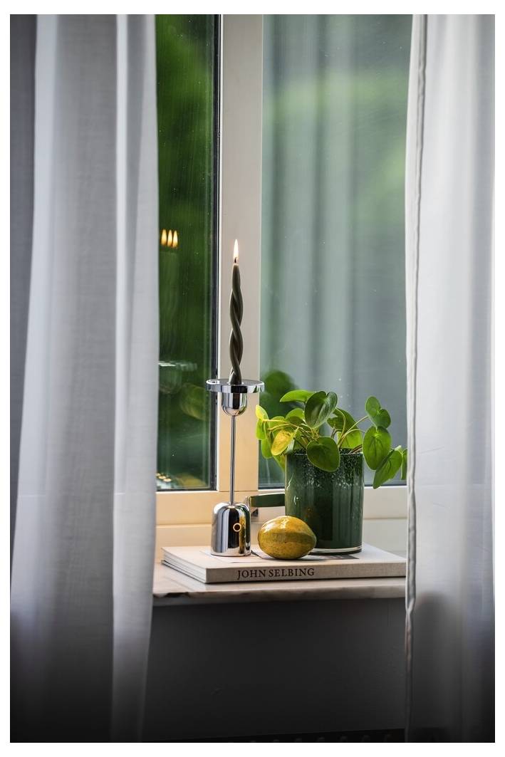 Un rebord de fenêtre décoré avec une bougie torsadée allumée, une plante verte en pot, un citron et des livres empilés, encadré par des rideaux légers.