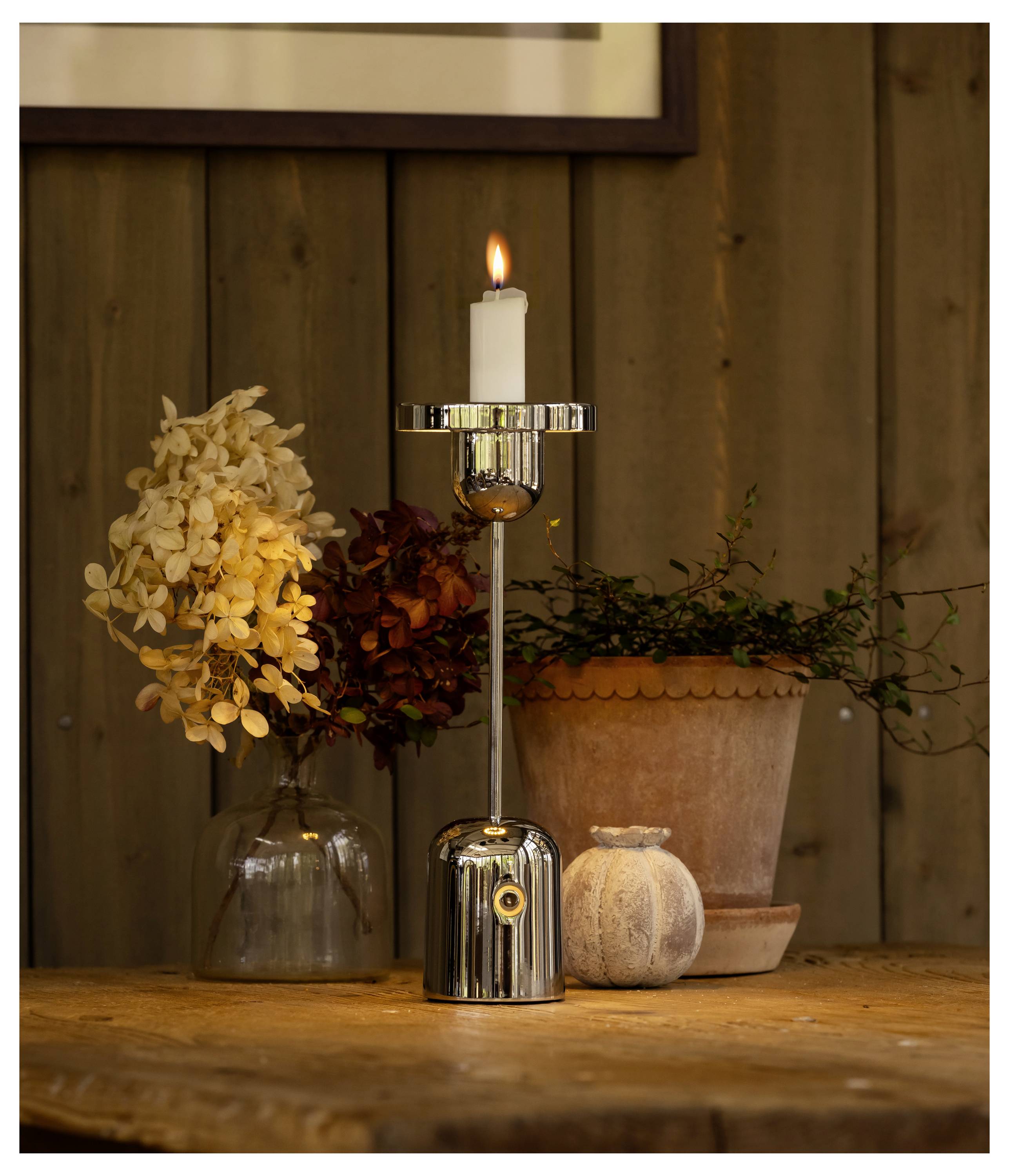 Une bougie allumée sur un support métallique posé sur une table en bois, accompagnée de fleurs séchées dans une bouteille en verre et d'un pot en argile, créant une ambiance chaleureuse.