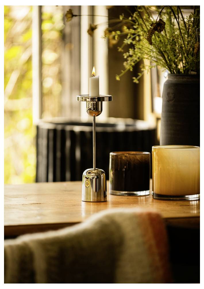 Une bougie allumée dans un support métallique sur une table en bois, encadrée par deux bougies dans des bocaux en verre et un vase garni de verdure dans une pièce chaleureuse.