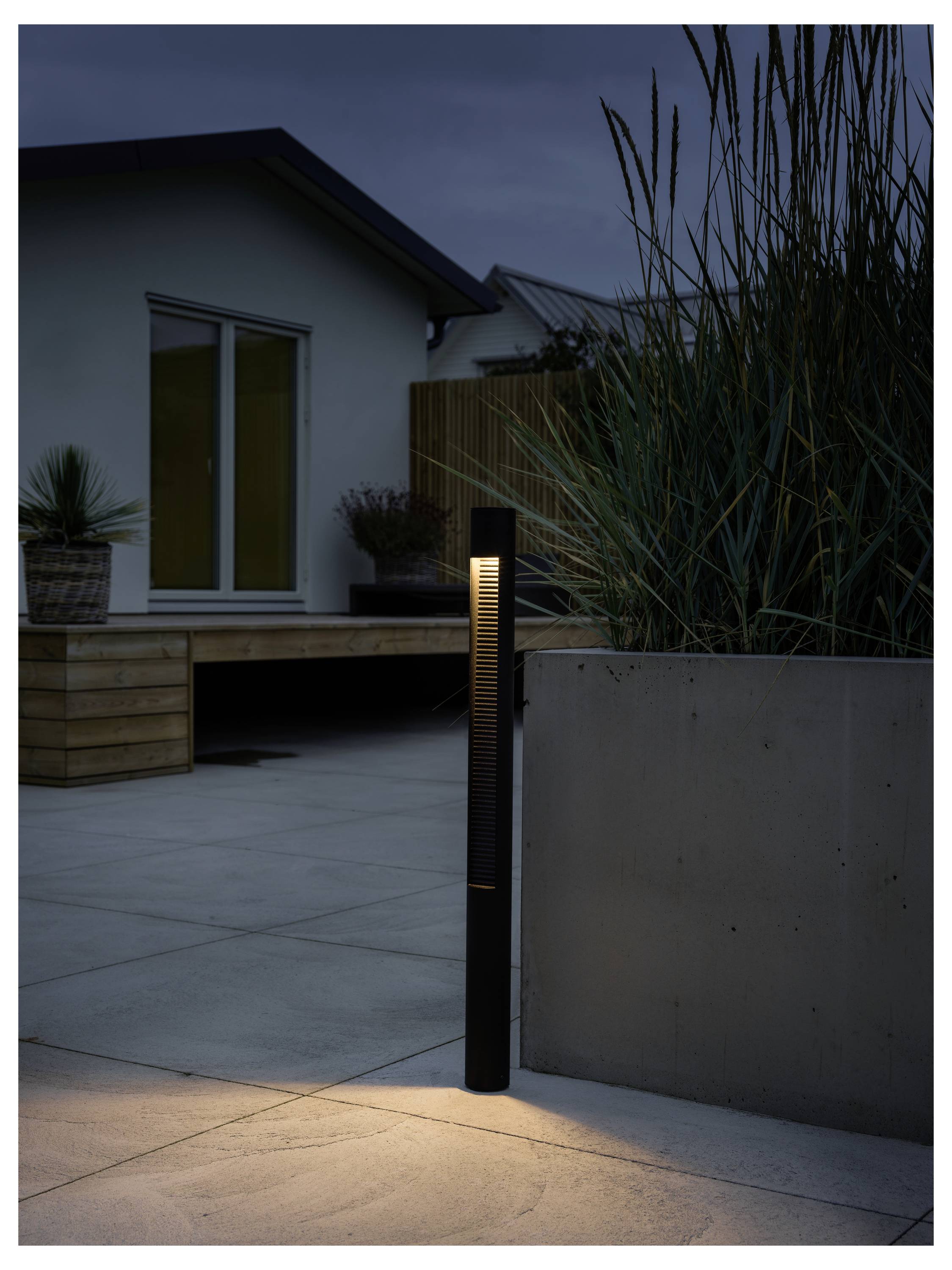 Une terrasse extérieure moderne au crépuscule avec des herbes hautes, un plancher en bois et un luminaire de sentier élégant projetant une lueur chaleureuse sur le sol.
