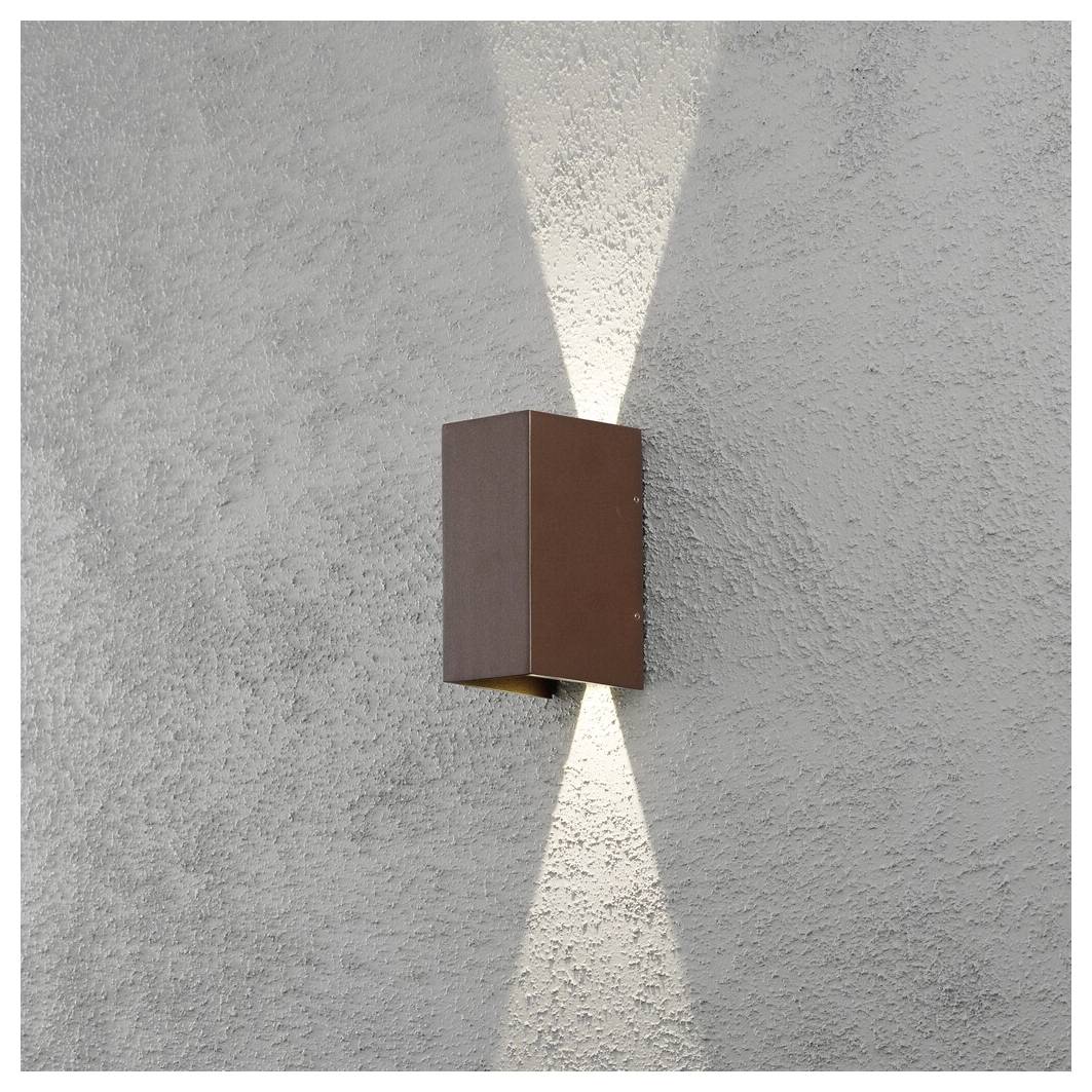 Un luminaire mural marron émet une lueur douce, à la fois vers le haut et vers le bas, contre un mur gris texturé, créant un motif symétrique.