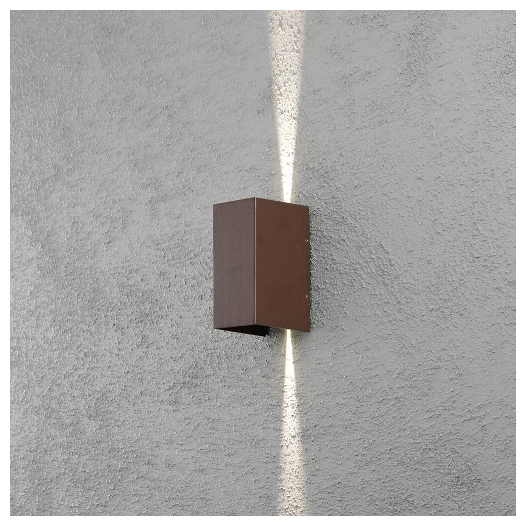 Un luminaire mural extérieur moderne, rectangulaire et marron, fixé sur un mur en stuc gris, émettant un éclairage vertical vers le haut et vers le bas.