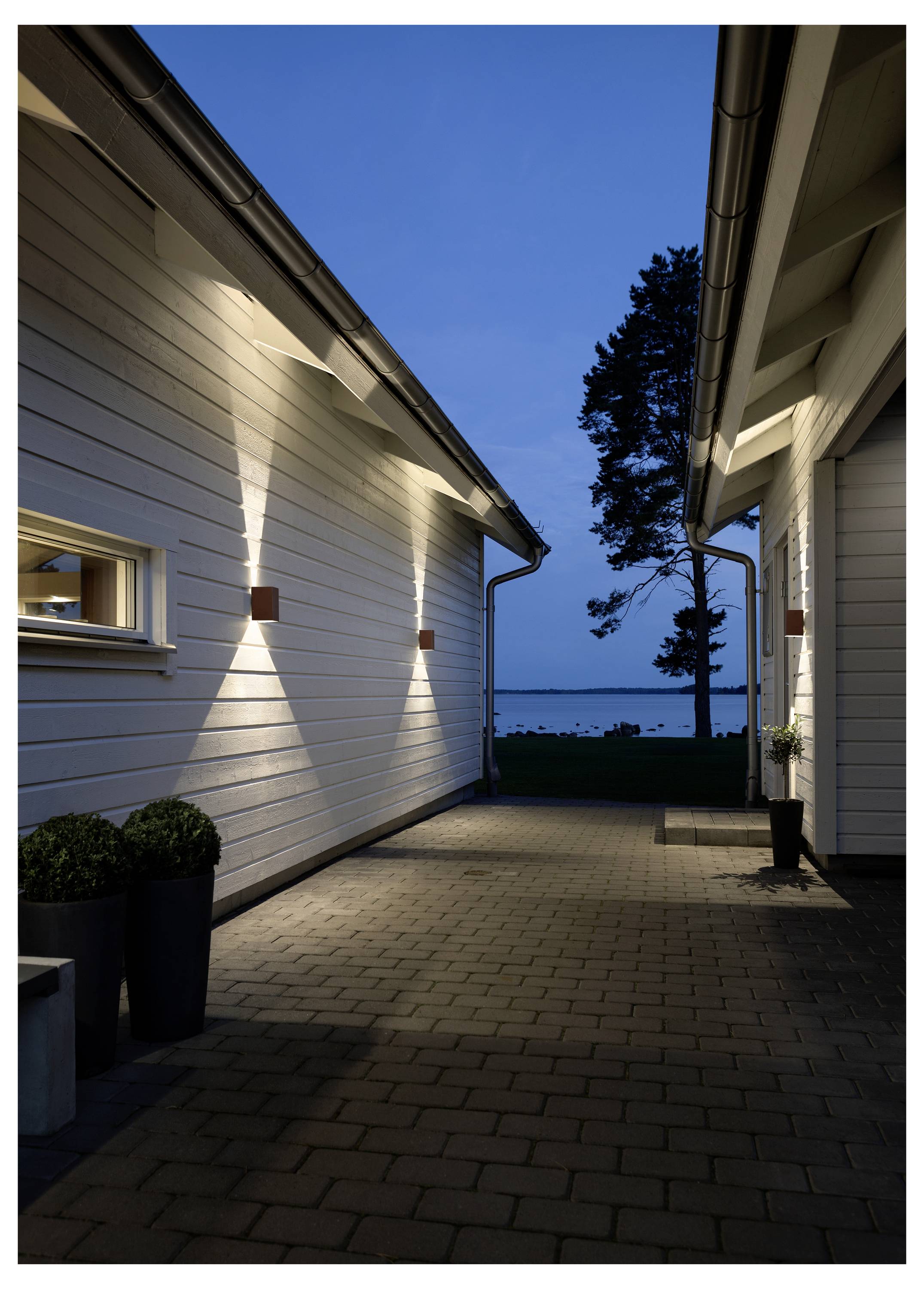 Une ruelle étroite entre deux maisons blanches modernes, avec des appliques murales projetant des faisceaux lumineux sur la façade, menant à une vue sur un lac au crépuscule.