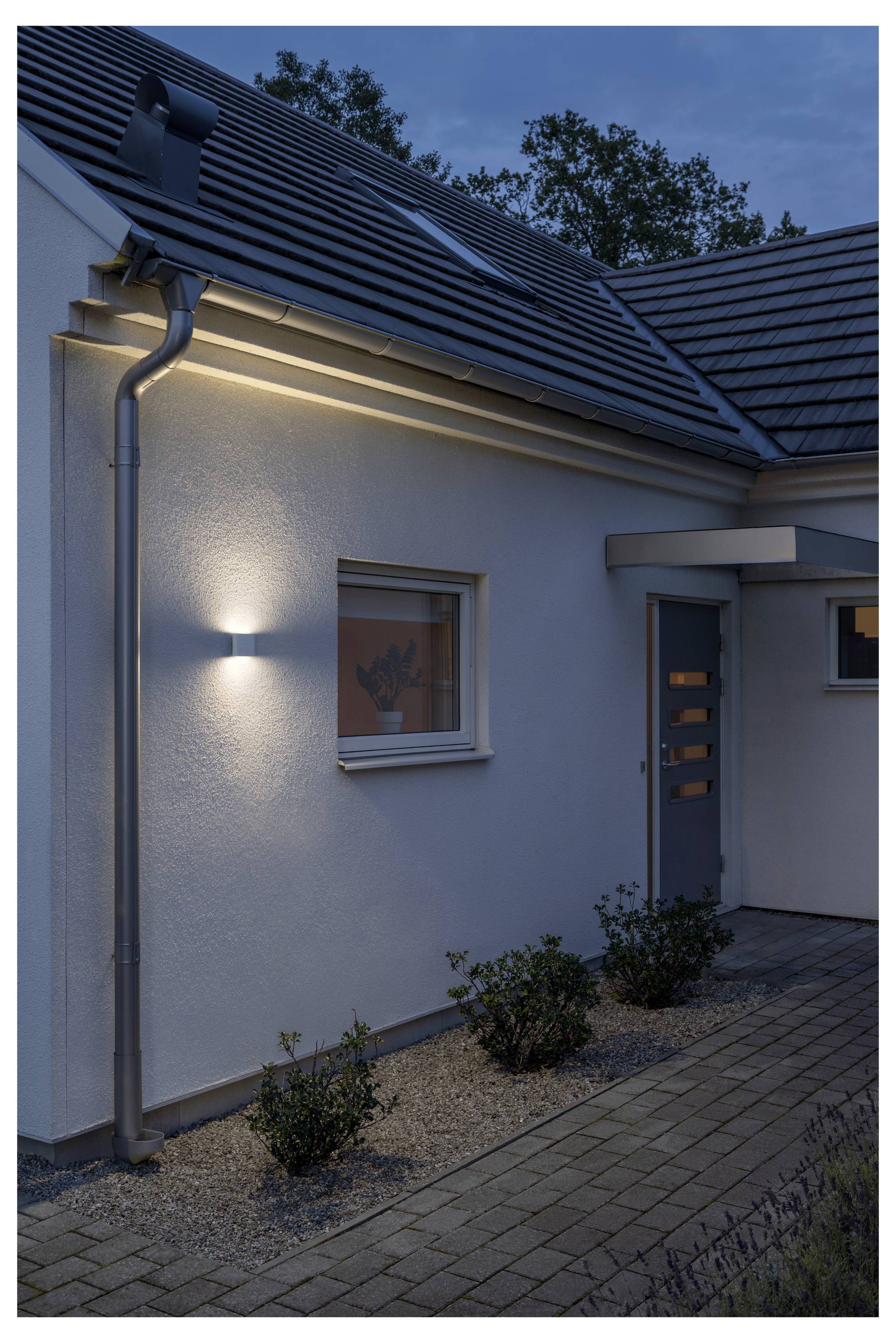 Extérieur d'une maison moderne au crépuscule avec un lampadaire extérieur illuminant une fenêtre et le chemin en contrebas.