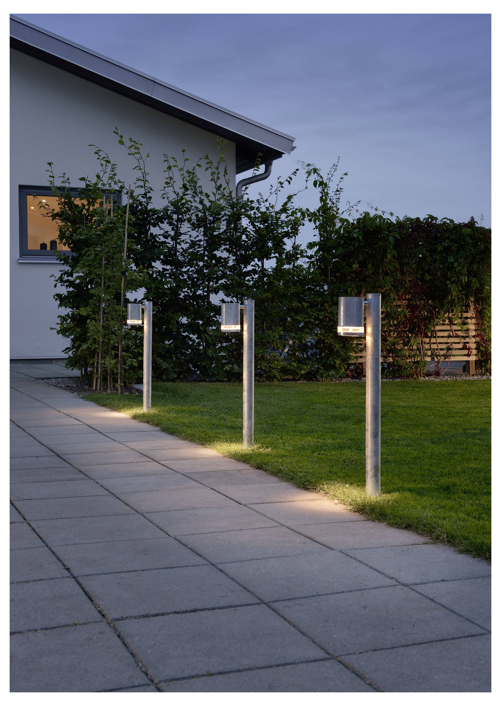 Un rang de lampes extérieures modernes illumine un chemin pavé à côté d'une maison, bordé d'herbe verte et de plantes pendant la soirée.