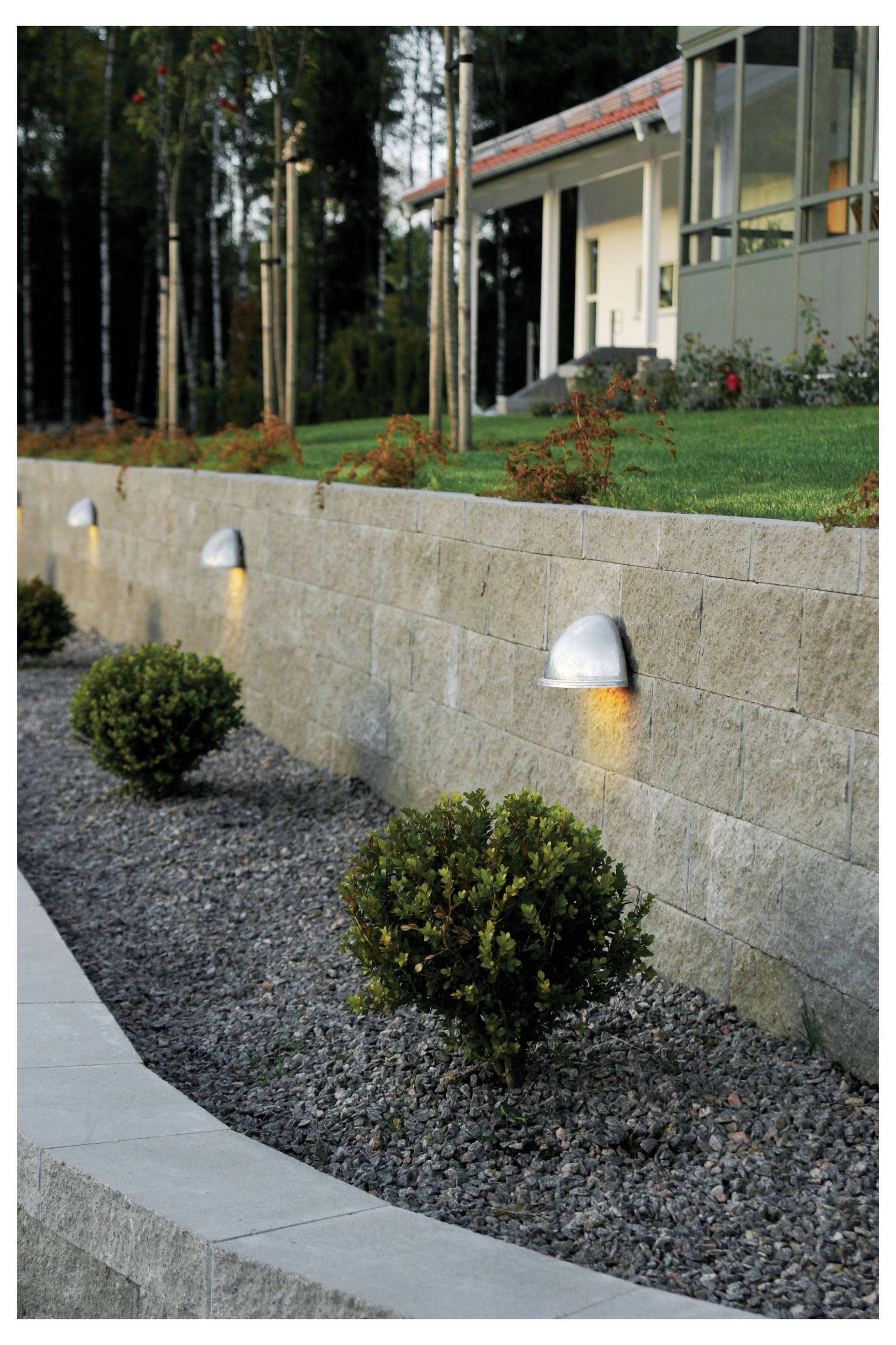 Une maison moderne avec une véranda vitrée, vue depuis un jardin aménagé avec des arbustes et un mur de soutènement en pierre, éclairé par des luminaires discrets au sol.