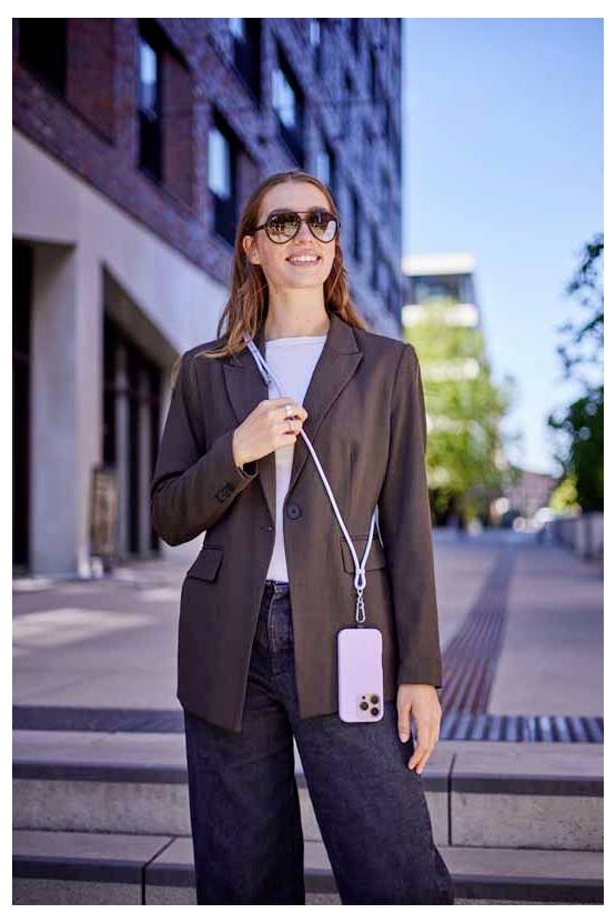 Une femme se tient dehors, souriante, portant des lunettes de soleil et une veste de tailleur, tenant un téléphone avec une lanière autour du cou.