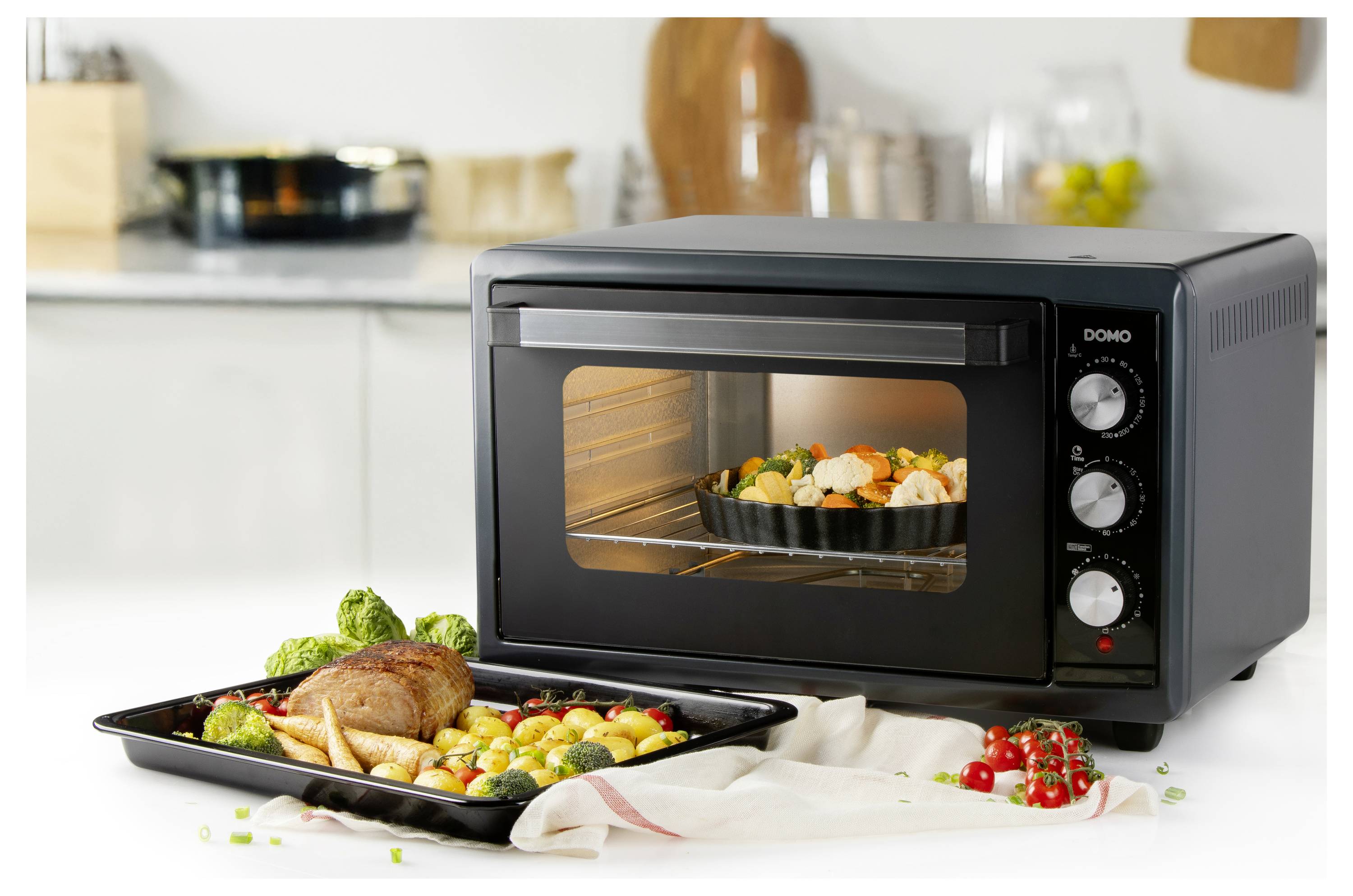 Un four de table dans une cuisine, en train de cuire un plat de légumes, avec un plateau de viande et de légumes rôtis placé devant. De la vaisselle de cuisine en arrière-plan.