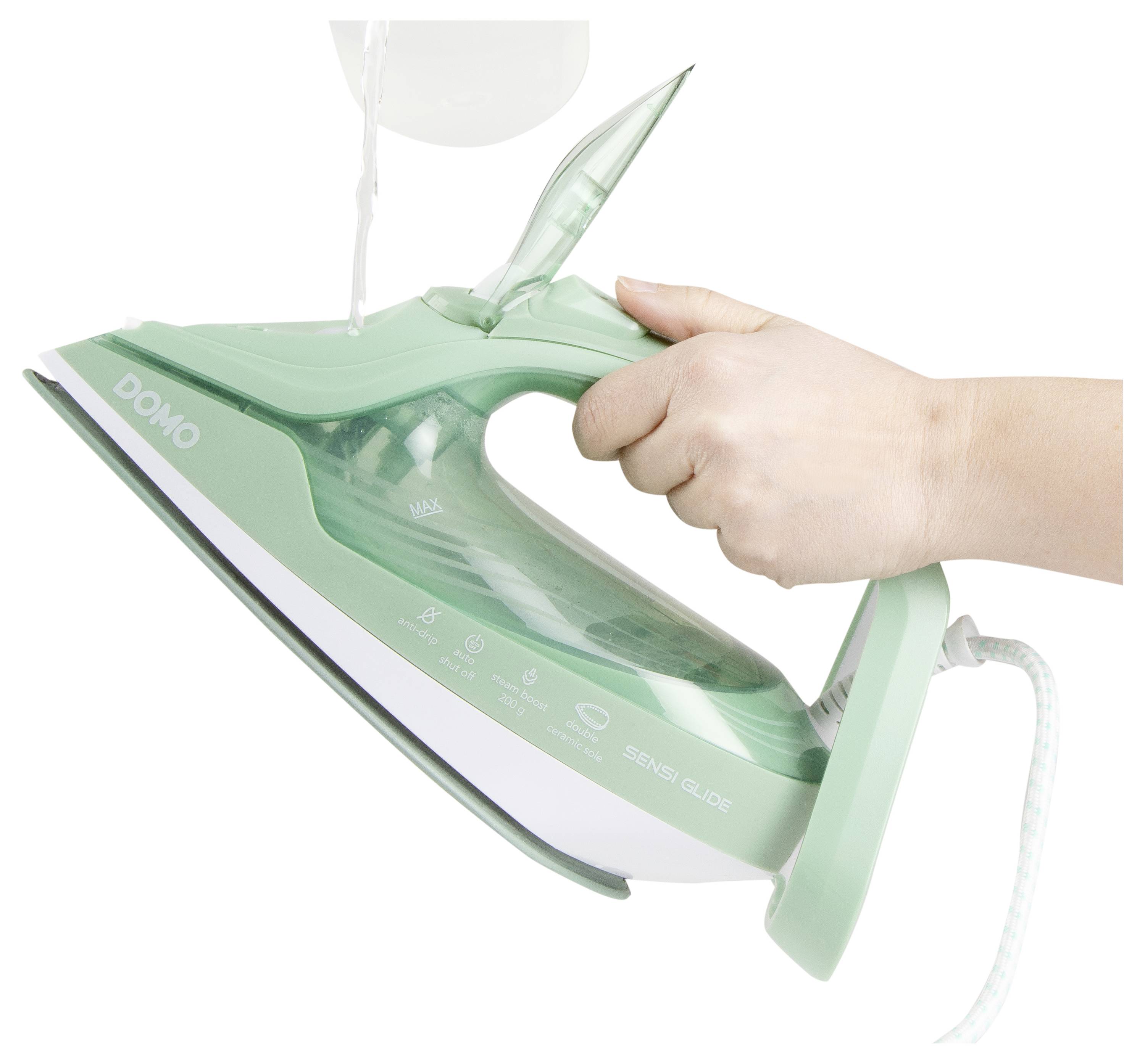 Une main tenant un fer à repasser vert portant l'étiquette 'DOMO', avec de l'eau versée dans son réservoir, prêt à être utilisé.