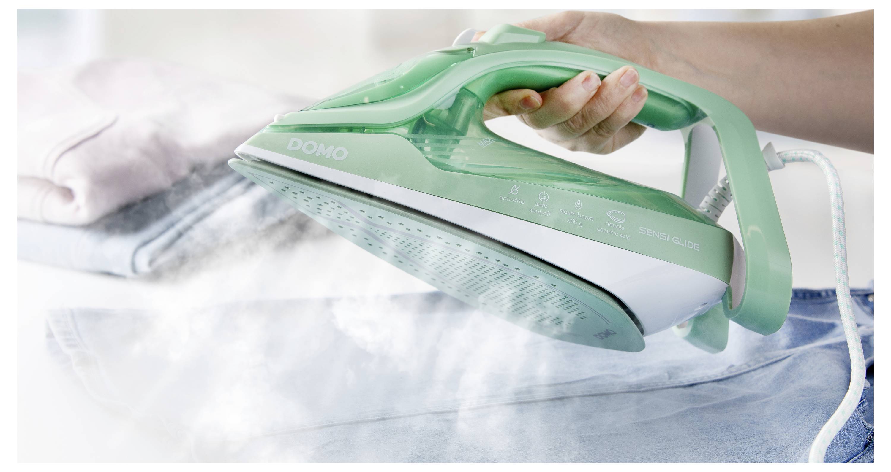 Une main tenant un fer à repasser à vapeur vert au-dessus d'un jean, libérant de la vapeur, avec des vêtements pliés en arrière-plan.