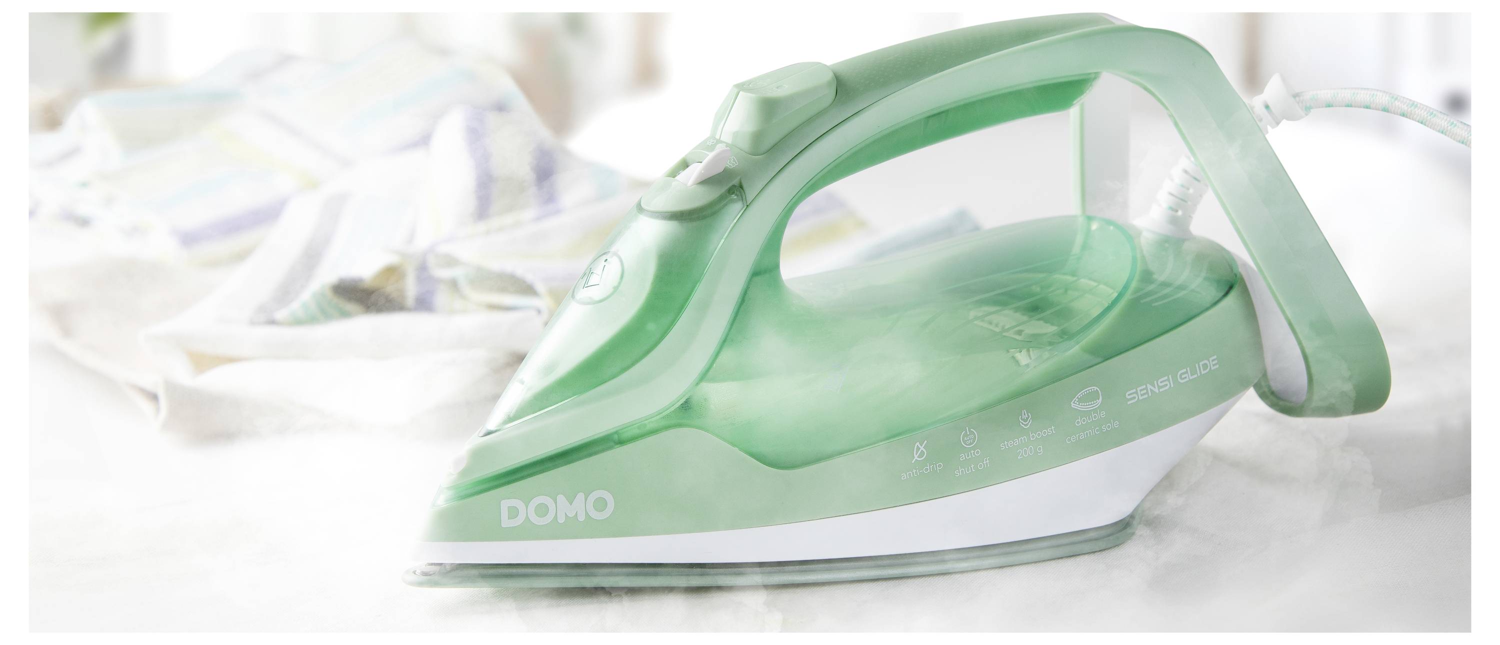 Un fer à repasser à vapeur vert et blanc portant l'étiquette 'DOMO' est posé sur un comptoir, avec une pile de tissus floue en arrière-plan.