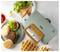 Un presse-sandwich vert avec une poignée en bois est entouré d'une assiette de steaks hachés, de pains à hamburger, de gaufres, de sandwichs grillés et de salade.