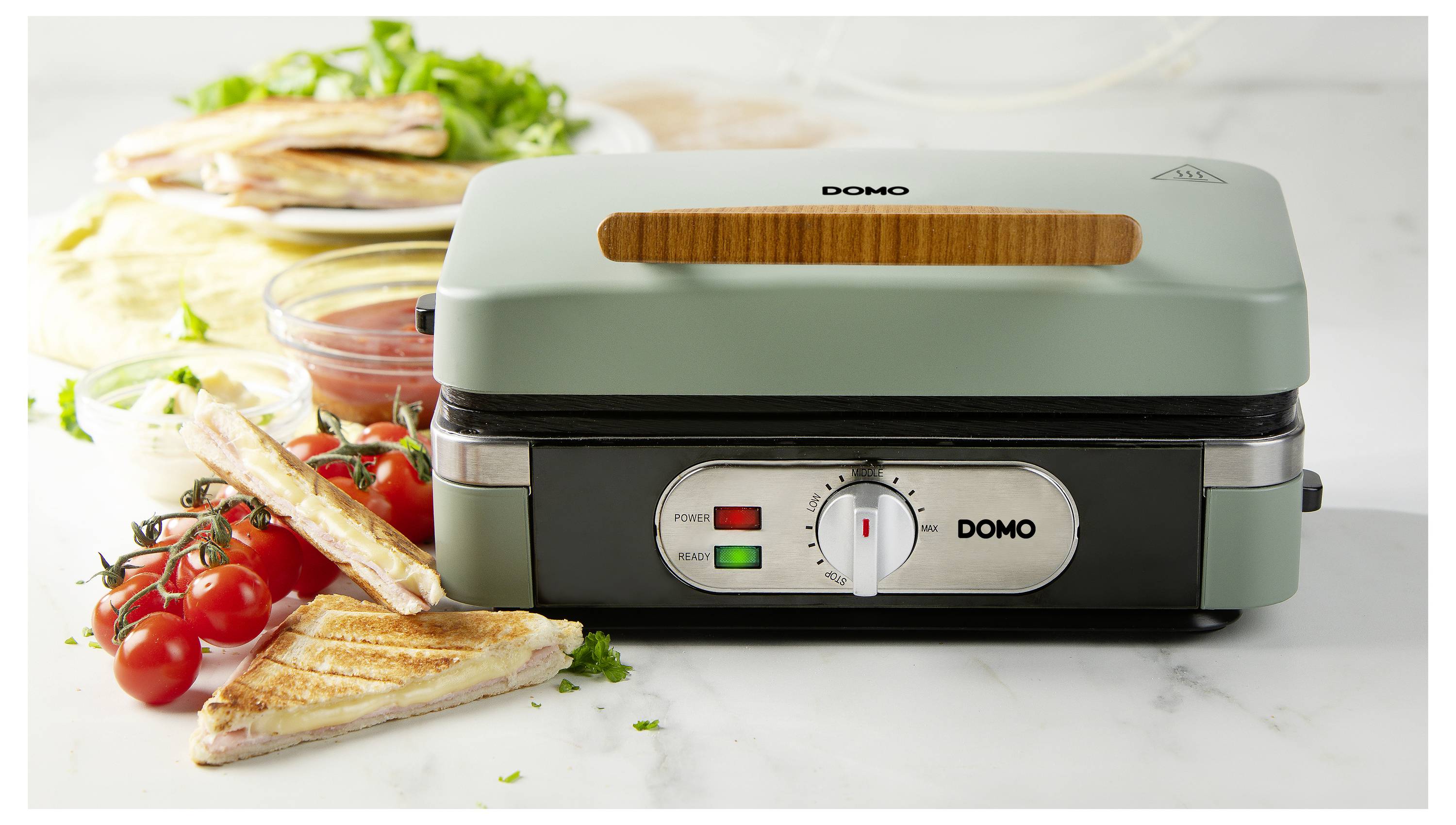 Un appareil à paninis est placé sur un plan de travail, entouré de sandwichs grillés et de tomates fraîches. Le voyant lumineux est allumé.