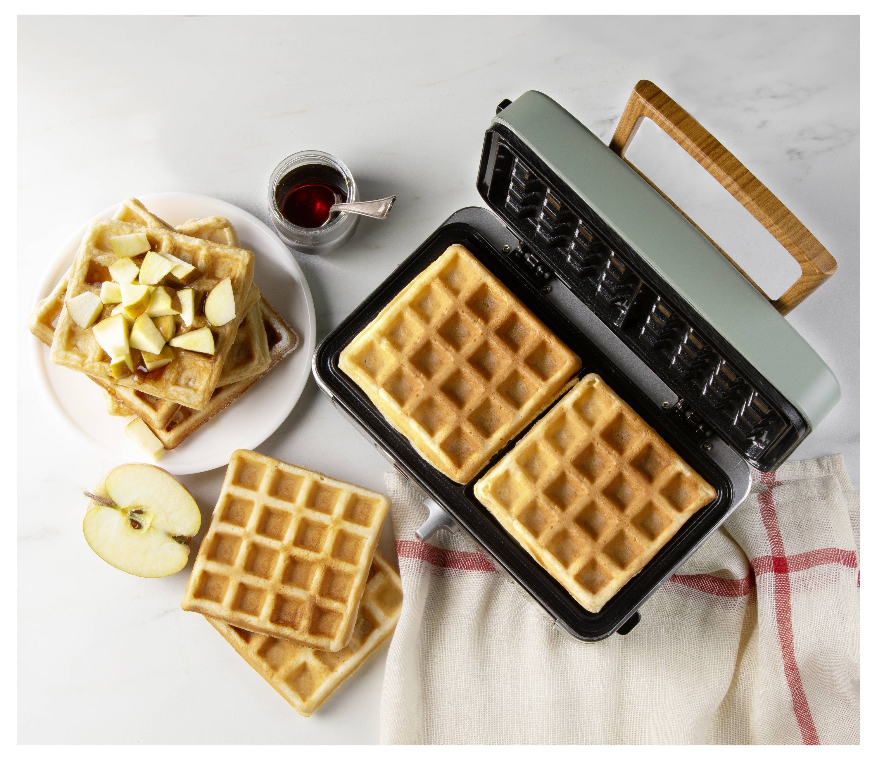 Un gaufrier avec deux gaufres cuites sur un plan de travail, entouré d'une assiette de gaufres avec des tranches de pommes, deux gaufres individuelles, du sirop et un torchon.