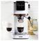 Machine à espresso versant du café dans une tasse en verre, avec un mousseur à lait attaché. Tasses et bocal à cookies en arrière-plan.