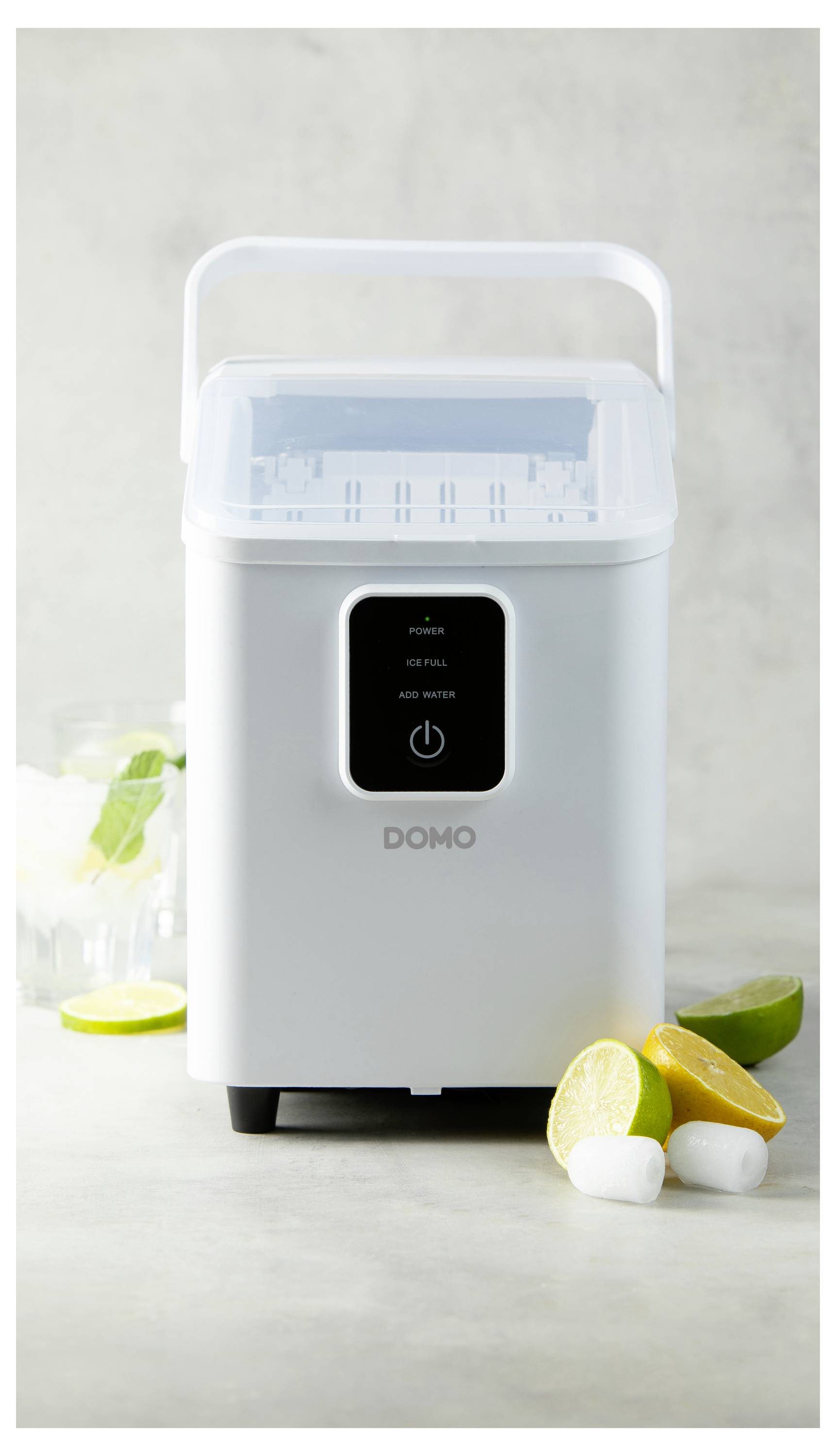Un fabricant de glaçons blanc est placé sur un plan de travail, entouré de glaçons et de citrons et limes coupés en deux.