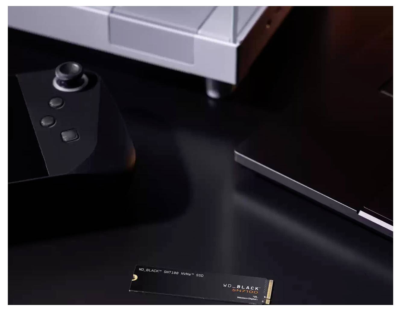 Un SSD NVMe WD_BLACK SN770 sur un bureau sombre, entouré d'une manette de jeu, d'un ordinateur portable et d'une partie d'un autre appareil électronique.