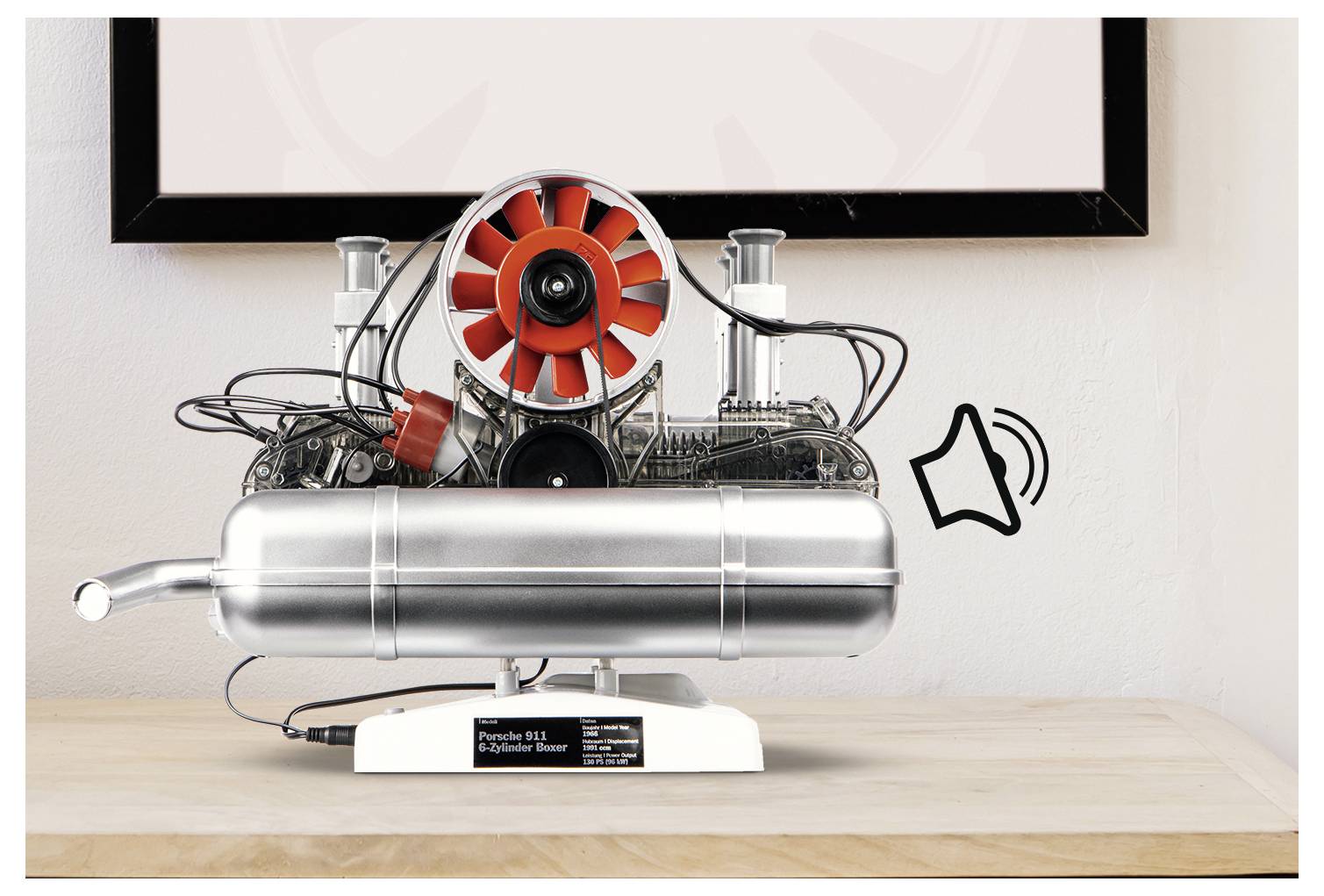 Un modèle détaillé d'un moteur de Porsche 911 posé sur une surface en bois, avec un ventilateur rouge et des câblages visibles. Une icône sonore suggère qu'il peut diffuser des sons de moteur.