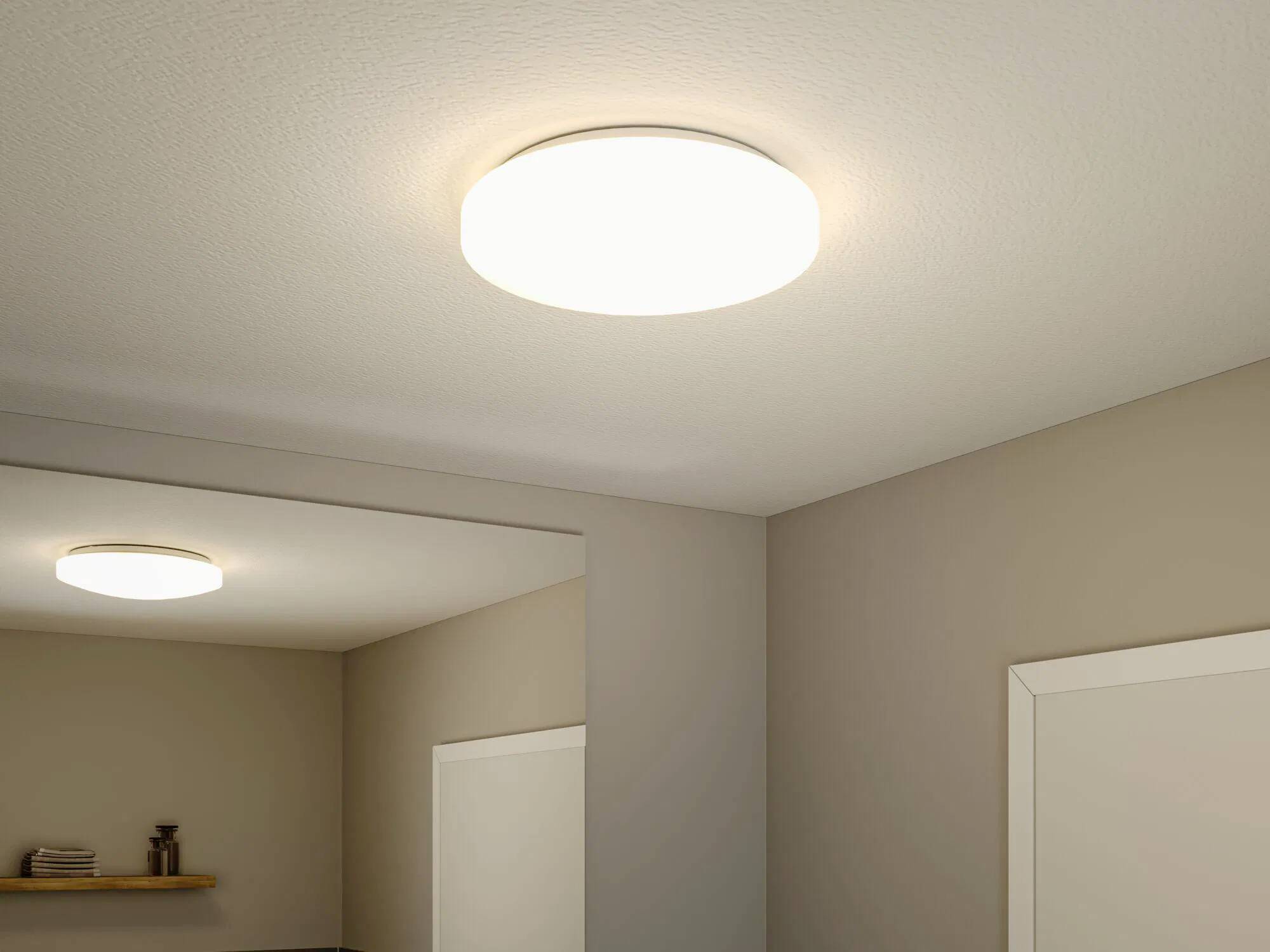 Plafond avec un luminaire LED rond illuminant une pièce aux tons neutres, avec un encadrement de porte et un miroir reflétant la lumière.